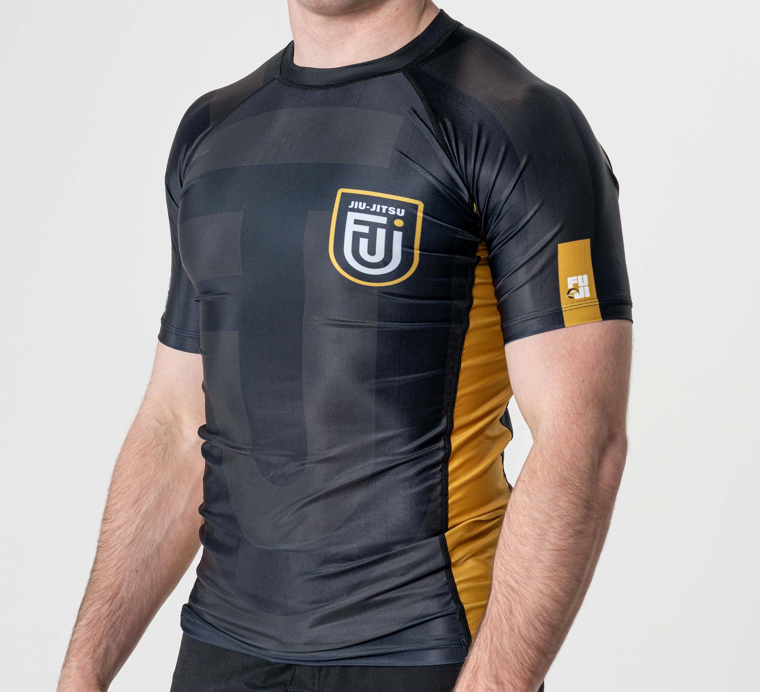 A person wearing the Rebel Flex Lite Rashguard Black with gold accents and FUJI Jiu-Jitsu logos on the chest and sleeve, standing against a plain white background.