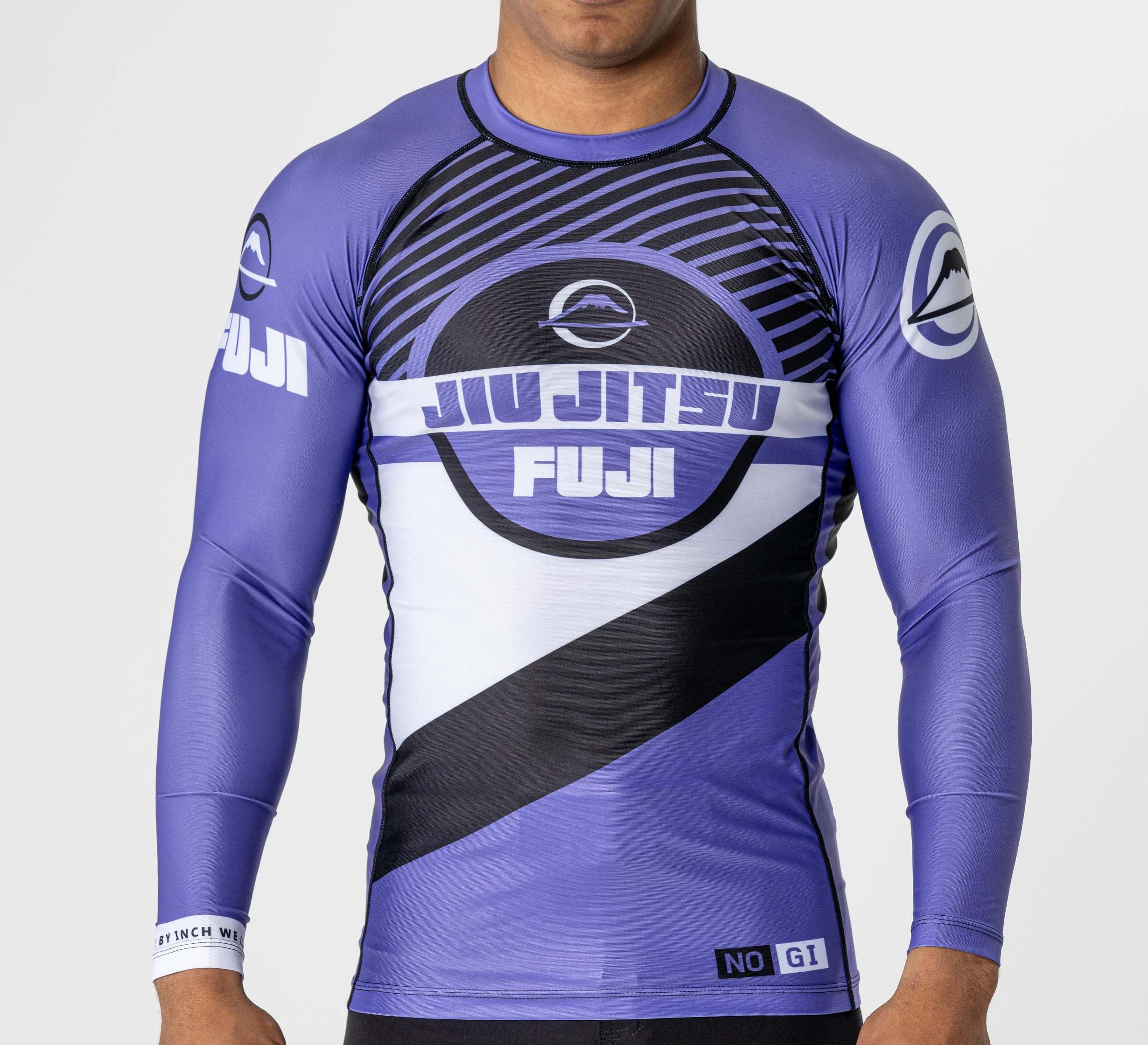 A person stands against a plain background wearing the FUJI Pacer Flex Lite Long Sleeve Rashguard in purple, featuring fitted sleeves and “JIU JITSU FUJI” text with graphic designs on the chest and arms.