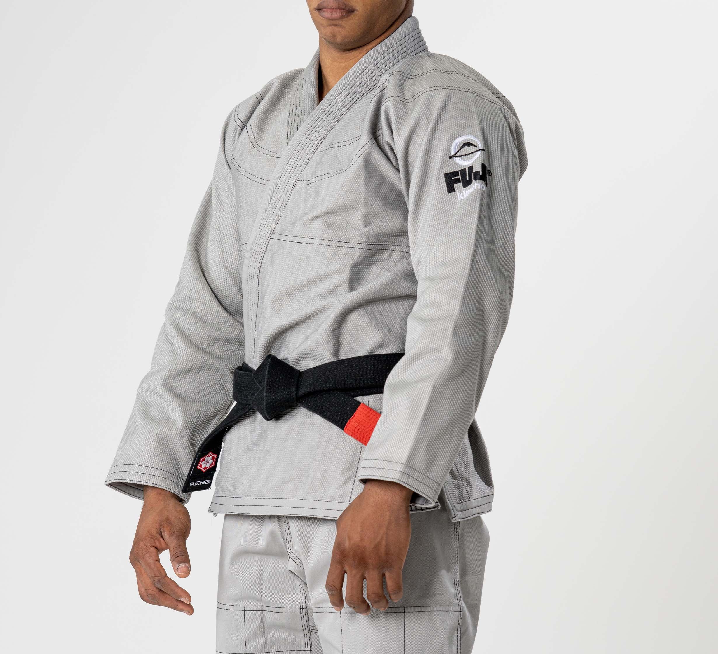 A person in the Lightweight BJJ Gi Grey with a black belt stands against a white background. The gi features a Fuji patch on the sleeve. The person's face is partially out of frame.