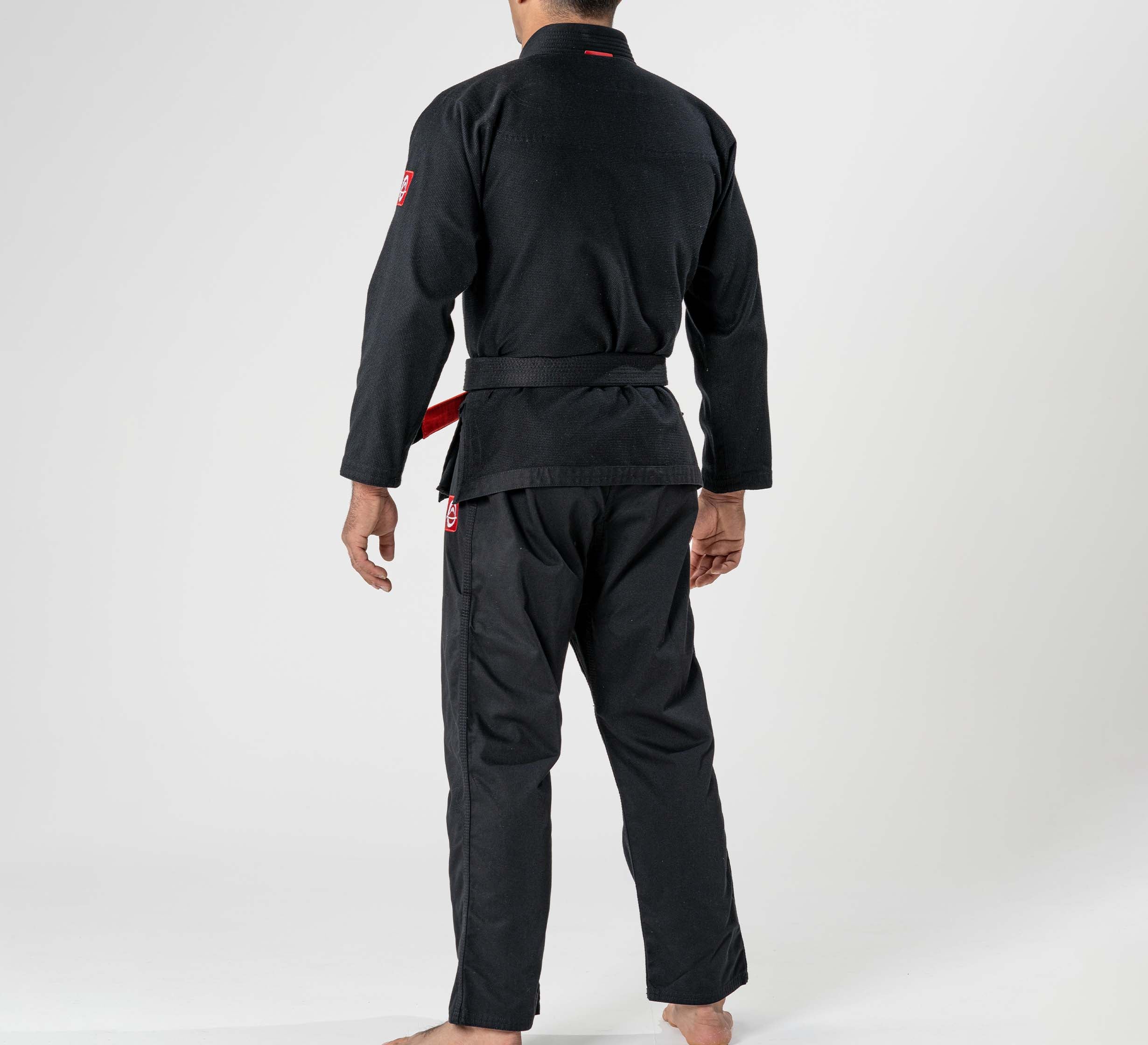 A person wearing the Ultra Lite 300 BJJ Gi Black with a black belt stands barefoot, facing away from the camera against a plain white background.