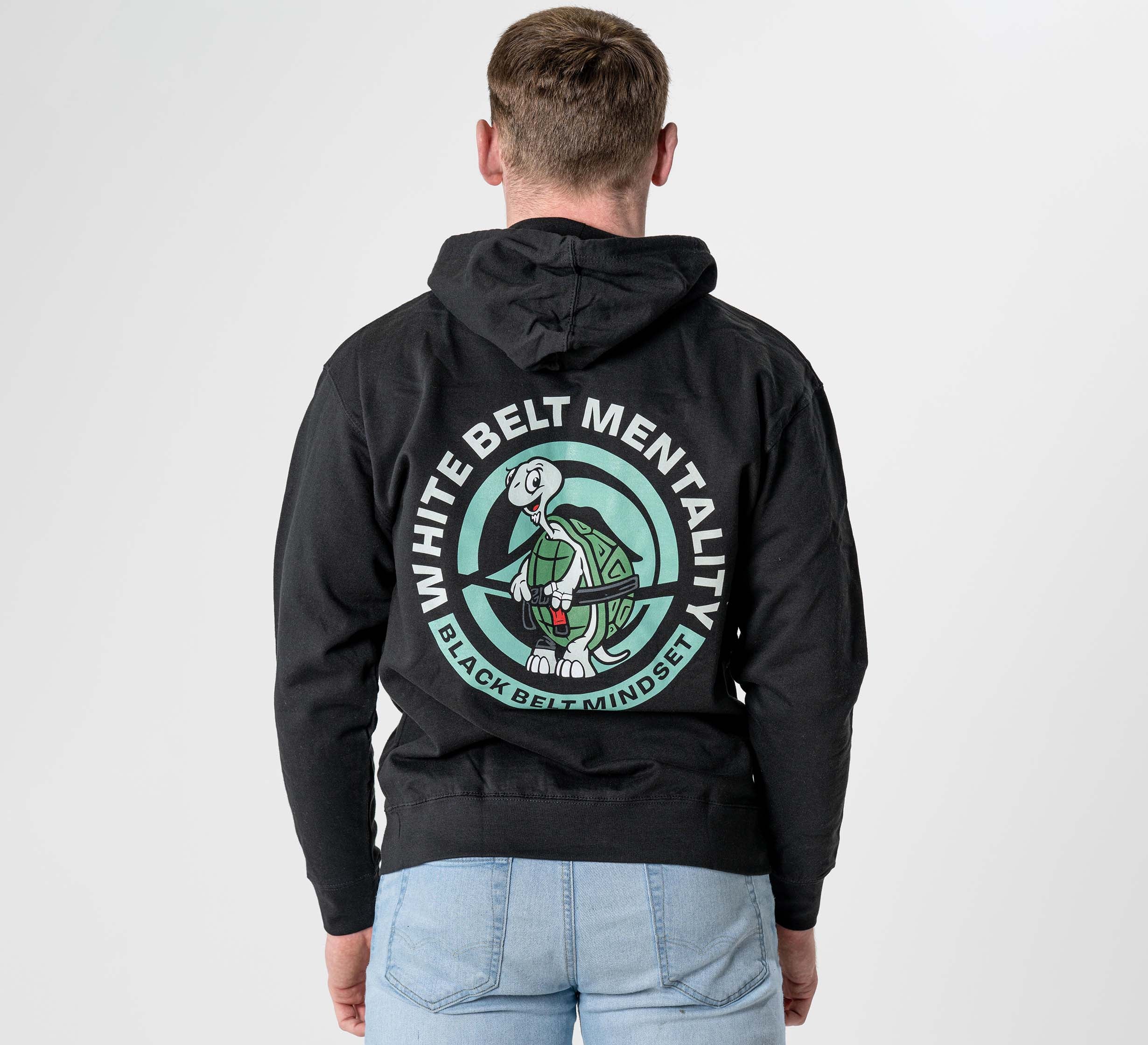 A person faces away, wearing the Black Belt Mentality Hoodie Black with a circular logo of a turtle holding a sword and the text "WHITE BELT MENTALITY" and "BLACK BELT MINDSET," paired with light blue jeans.