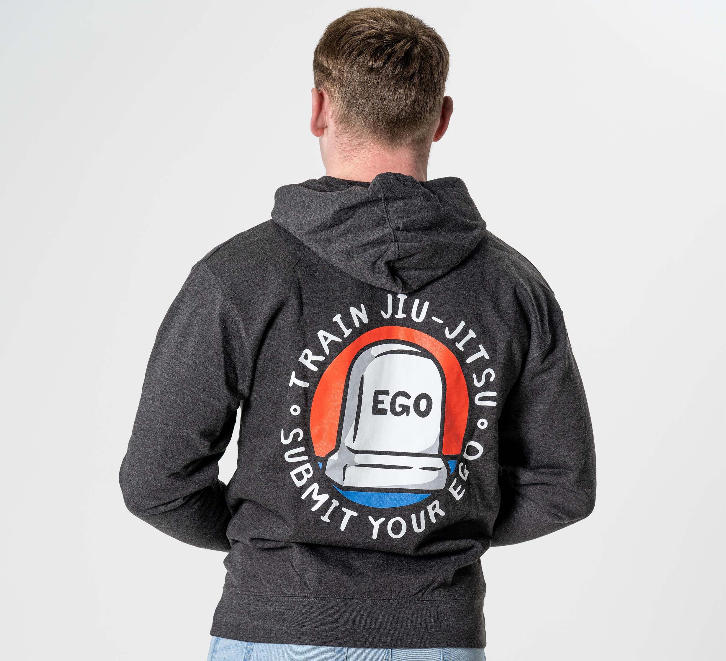 Someone faces away from the camera, wearing the Submit Your Ego Hoodie in Heather Black, featuring a tombstone graphic labeled "EGO" and the words "Train Jiu-Jitsu, Submit Your Ego" in a circle on the back.