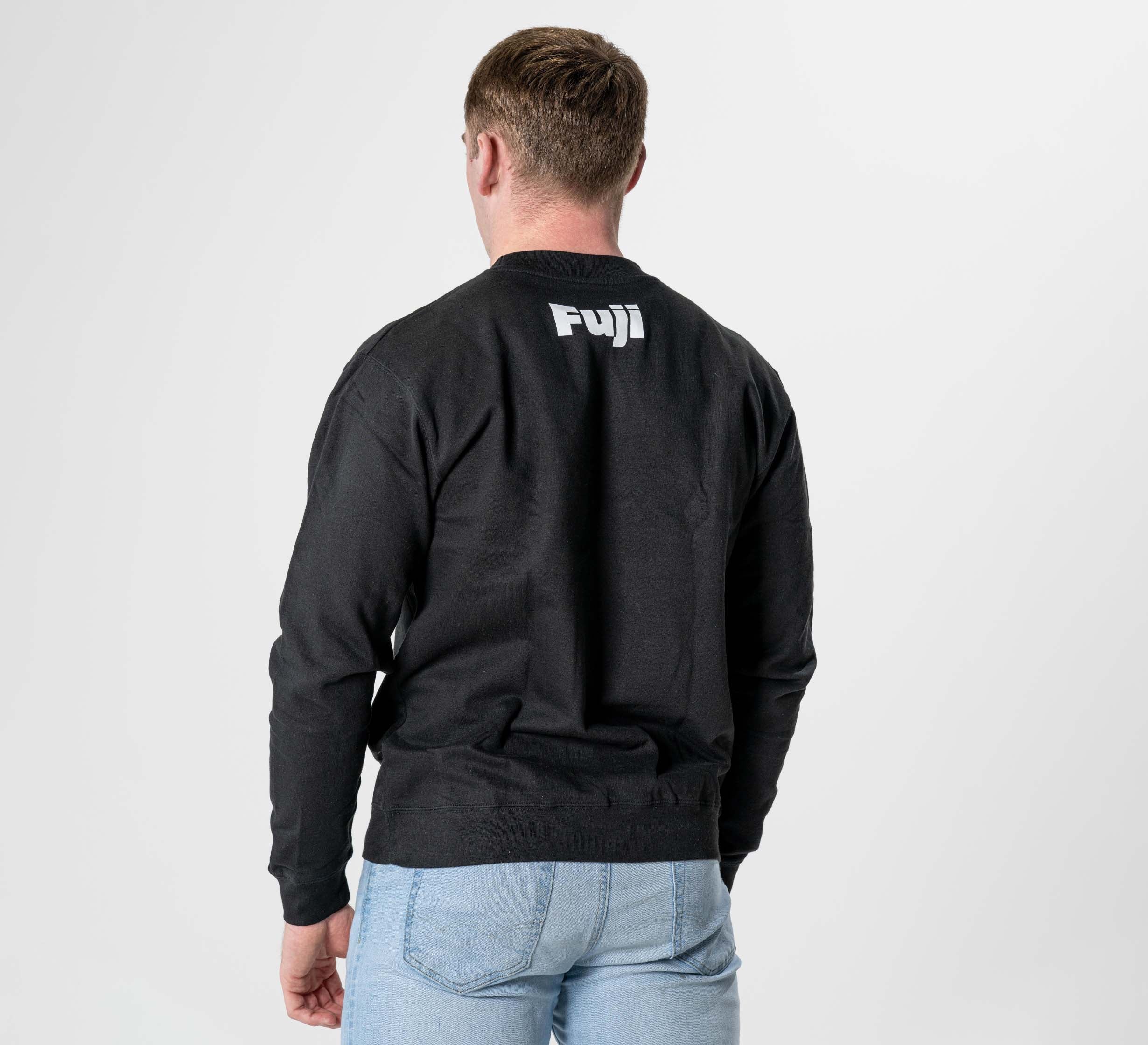 A person stands with their back to the camera, wearing the Jiu Jitsu Player Crewneck Black featuring “Fuji” in white on the upper back, paired with light blue jeans against a plain white background.
