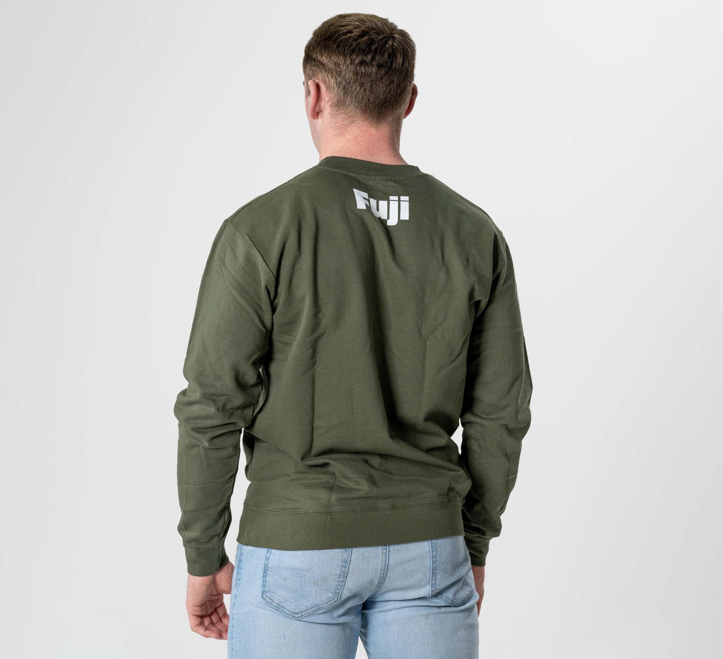 A man stands facing away against a plain white background, wearing the Jiu Jitsu Player Crewneck in military green with the white Fuji logo on the back, paired with light blue jeans.