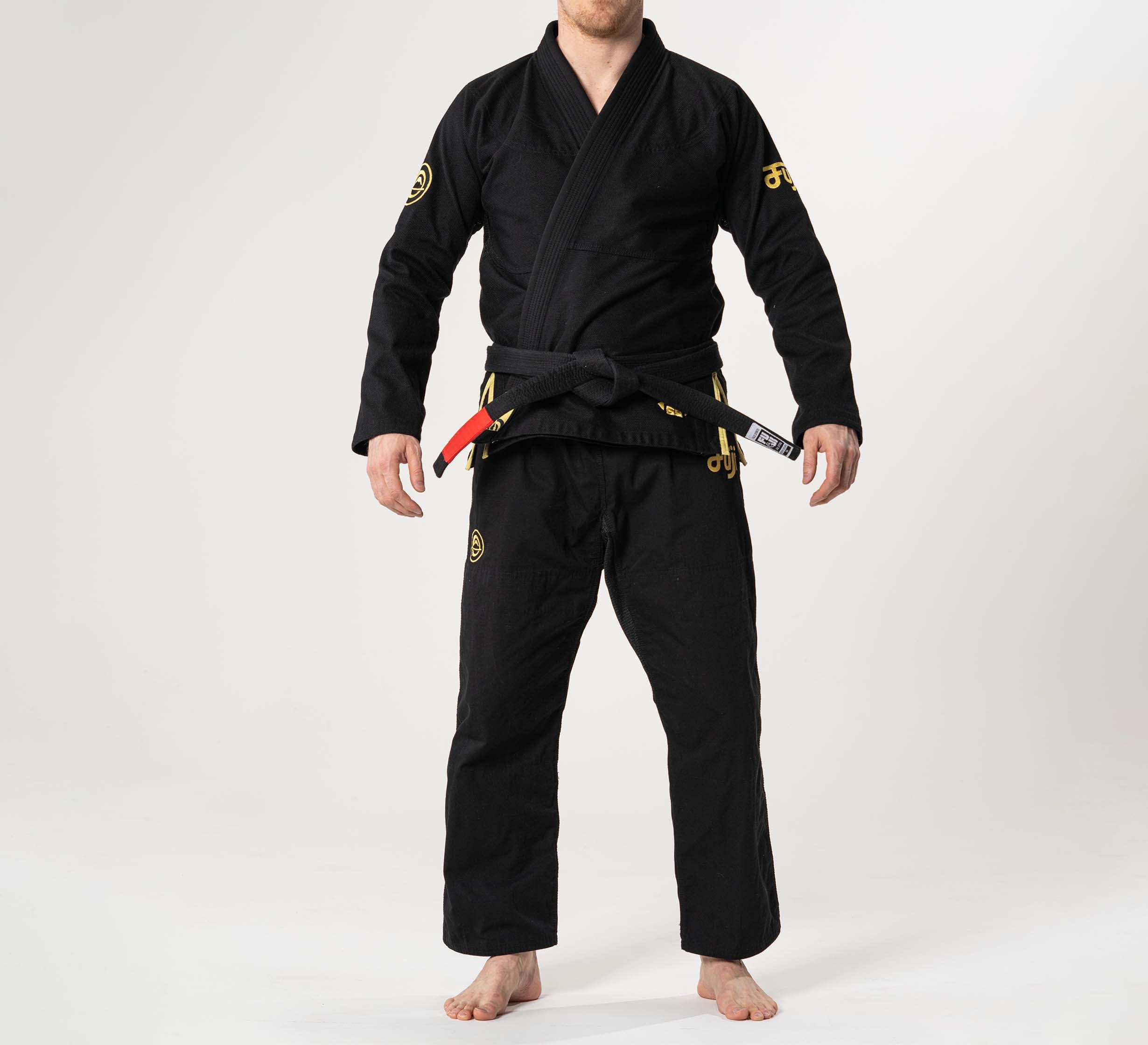 A person stands barefoot on a white background, wearing the Flow-Tech BJJ Gi Black/Gold with yellow patches and a black belt featuring red and white rank tags. Their face is not visible.