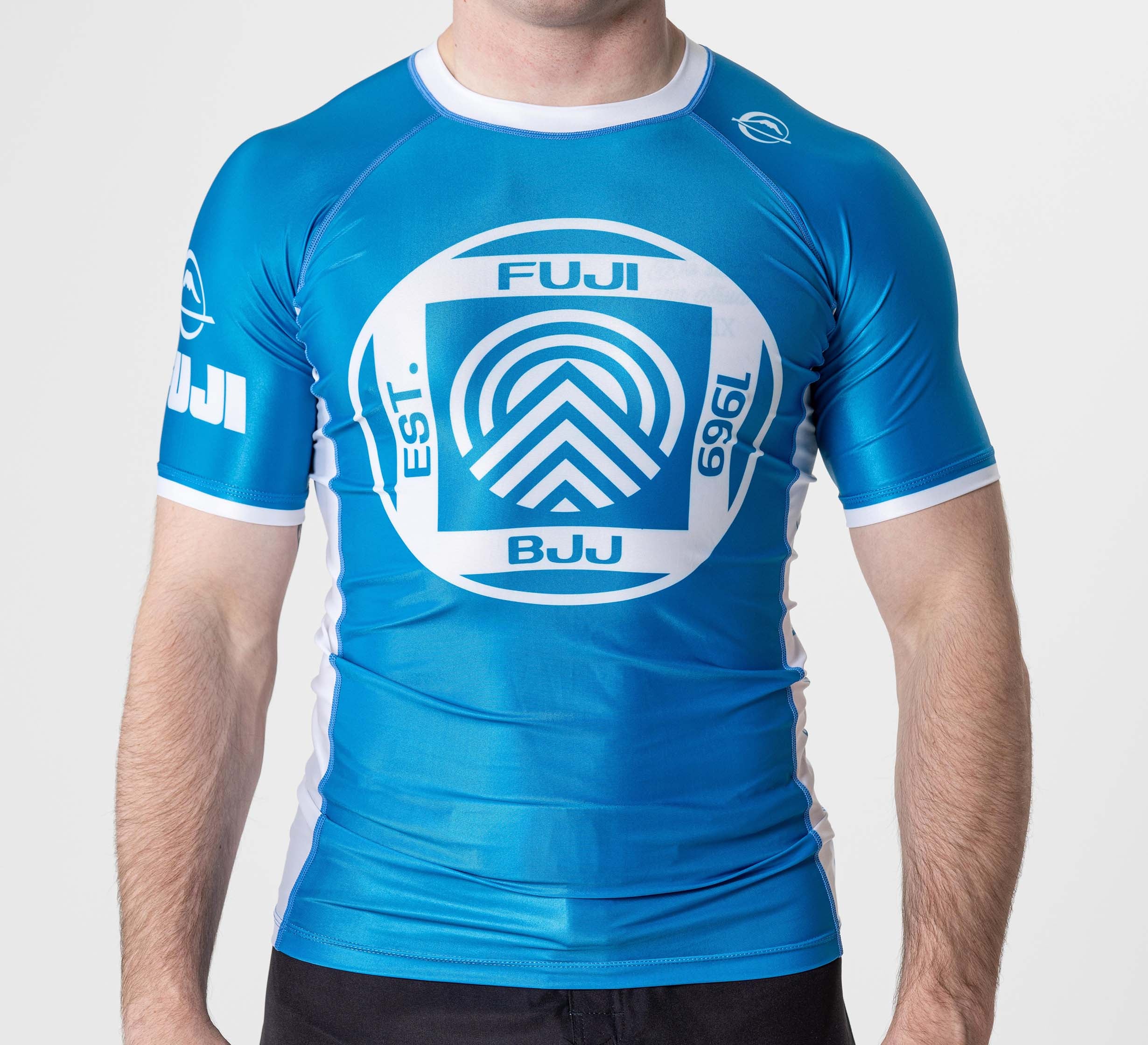 A person wearing the BJJ Flex Lite Rashguard Blue, a blue and white short-sleeve athletic top with "FUJI BJJ EST. 1969" and a circular logo on the chest. The person's head is not shown.