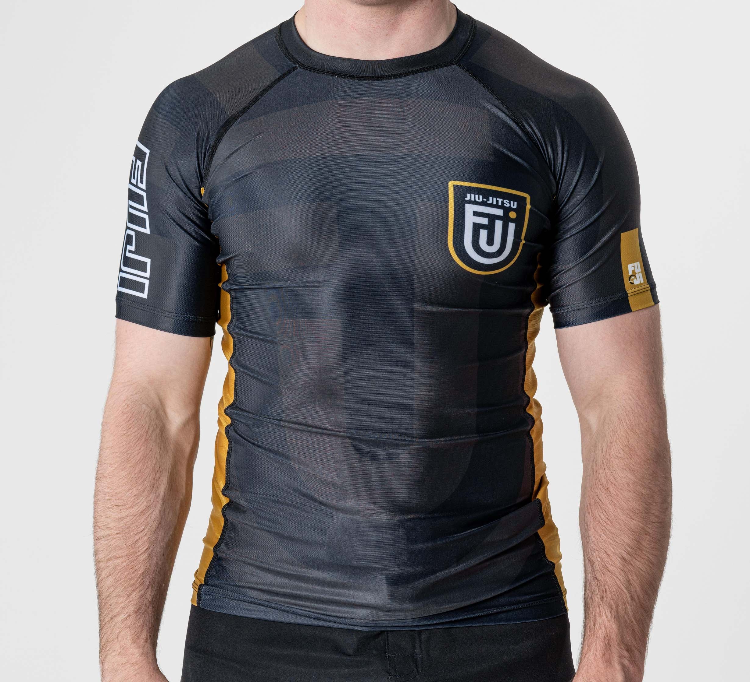 Someone wearing the Rebel Flex Lite Rashguard Black, a tight black rash guard with yellow accents, Jiu-Jitsu and a stylized JJ logo on the chest, plus white text on the left sleeve, stands against a plain background.