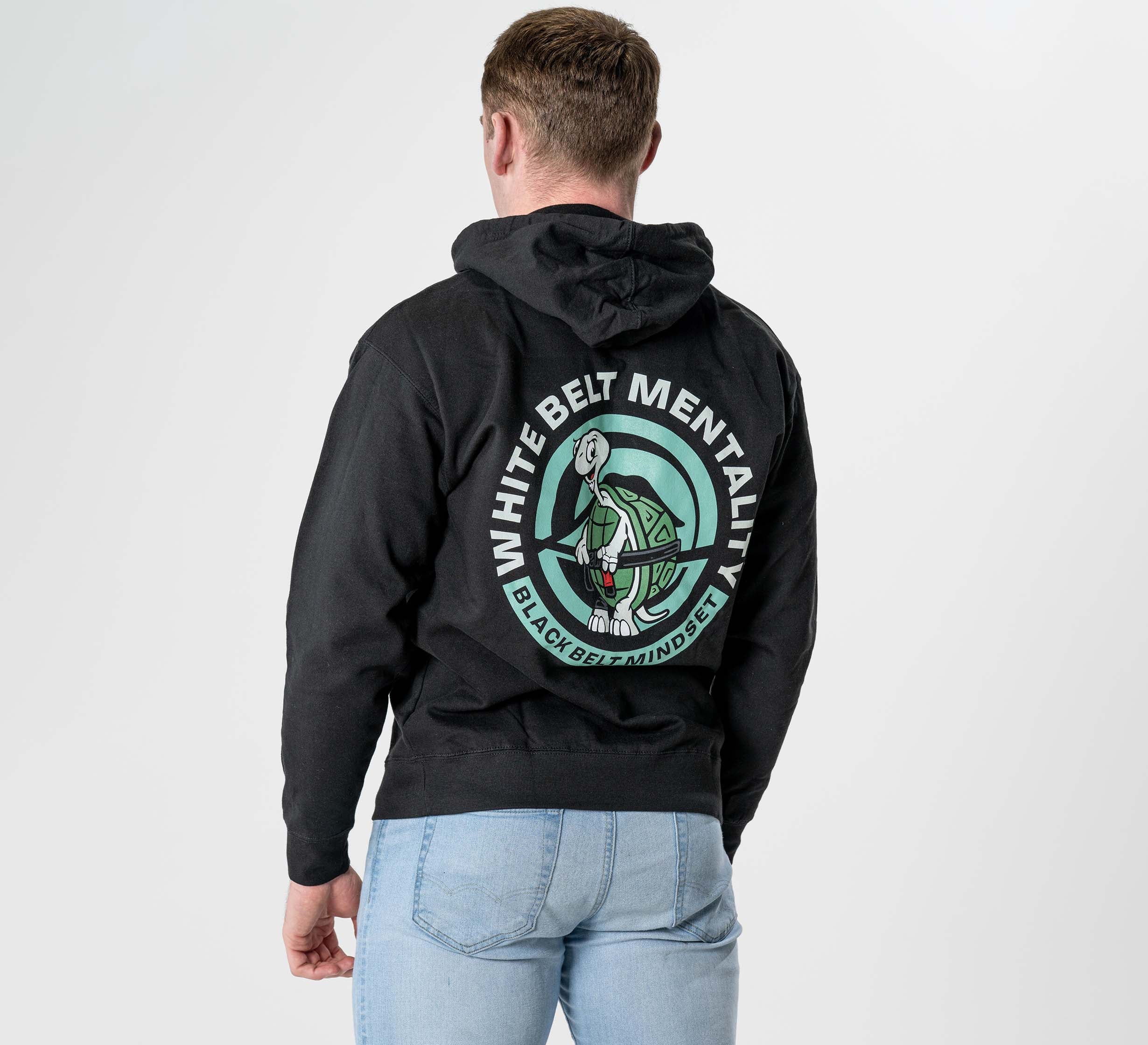 Someone wearing the Black Belt Mentality Hoodie Black, featuring a martial artist graphic and "White Belt Mentality, Black Belt Training Dept" on the back, paired with light blue jeans, stands facing away from the camera.
