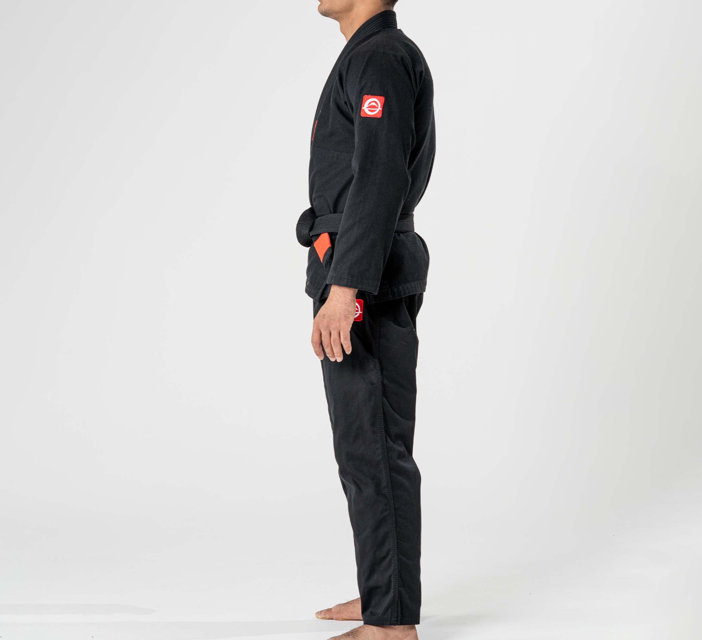 A child stands barefoot in profile view wearing the Kids Ultra Lite 300 BJJ Gi Black, featuring red sleeve patches, against a plain white background.