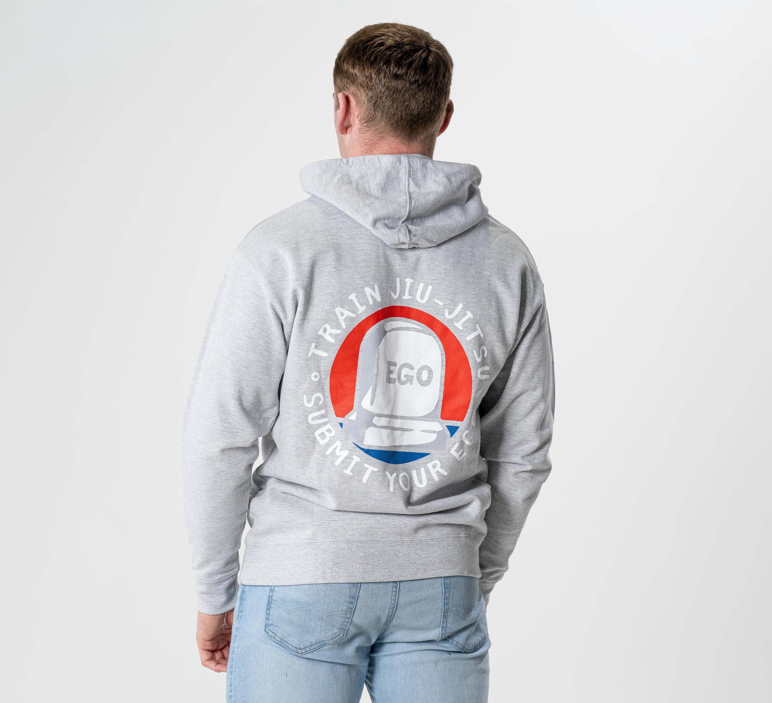 A person in a heather grey "Submit Your Ego Hoodie" is seen from behind against a plain light background. The hoodie features a tombstone graphic with “EGO” and the circular phrase “TRAIN JIU-JITSU SUBMIT YOUR EGO.”.