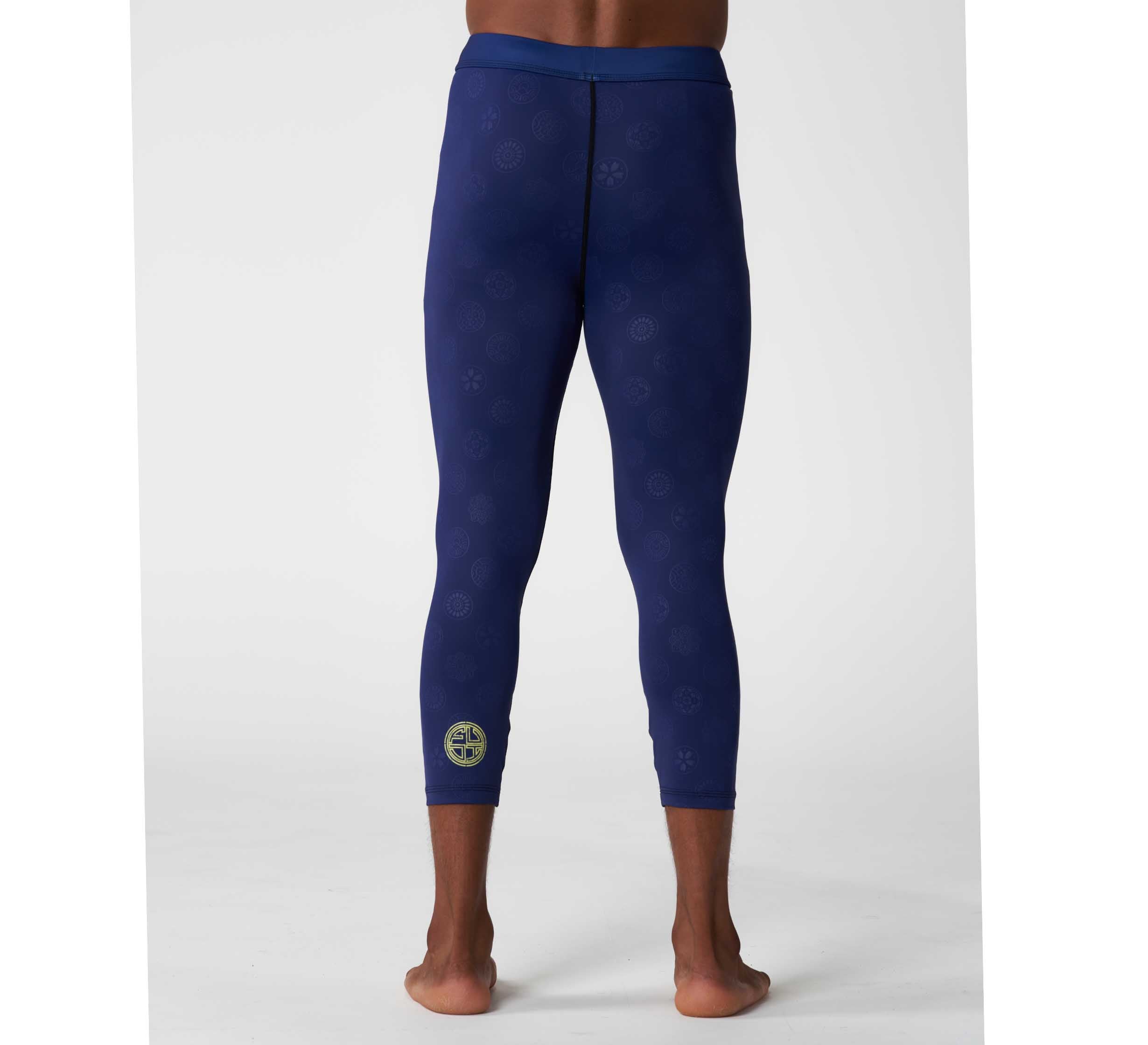 A person stands barefoot, facing away, wearing Shogun Heat Gear Spats Navy—dark blue fitted athletic leggings with a subtle pattern and a small yellow circular logo on the lower left leg. The background is plain white.