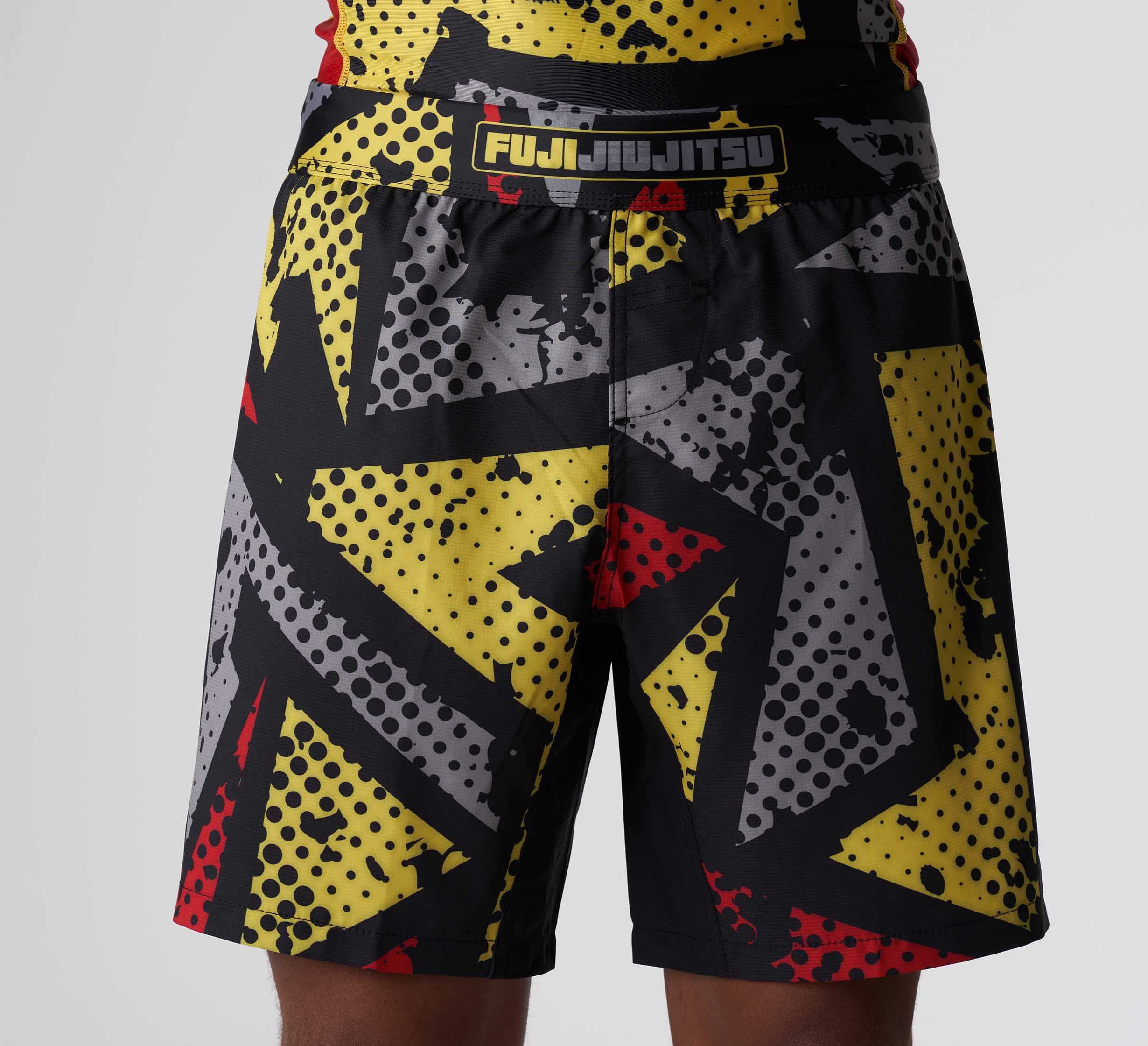 Person wearing Shuriken Flex Lite Shorts Black/Yellow, featuring a bold graphic design with black, yellow, red, and gray jagged and dotted patterns. The waistband displays the FUJI JIU JITSU logo in yellow and red.