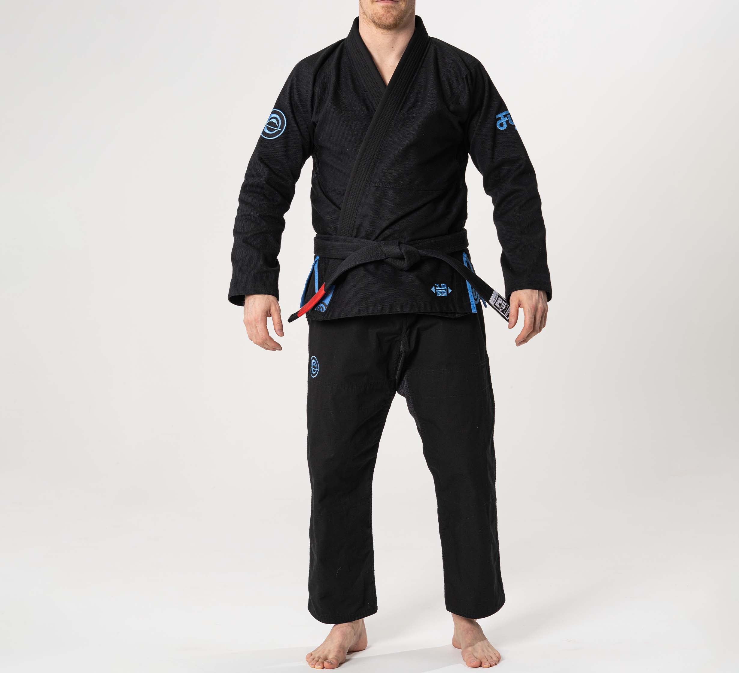 A person stands barefoot on a white background wearing the Flow-Tech BJJ Gi in black with blue and grey accents, featuring a black belt with red and blue rank stripes. Their hands are relaxed at their sides, head slightly turned.