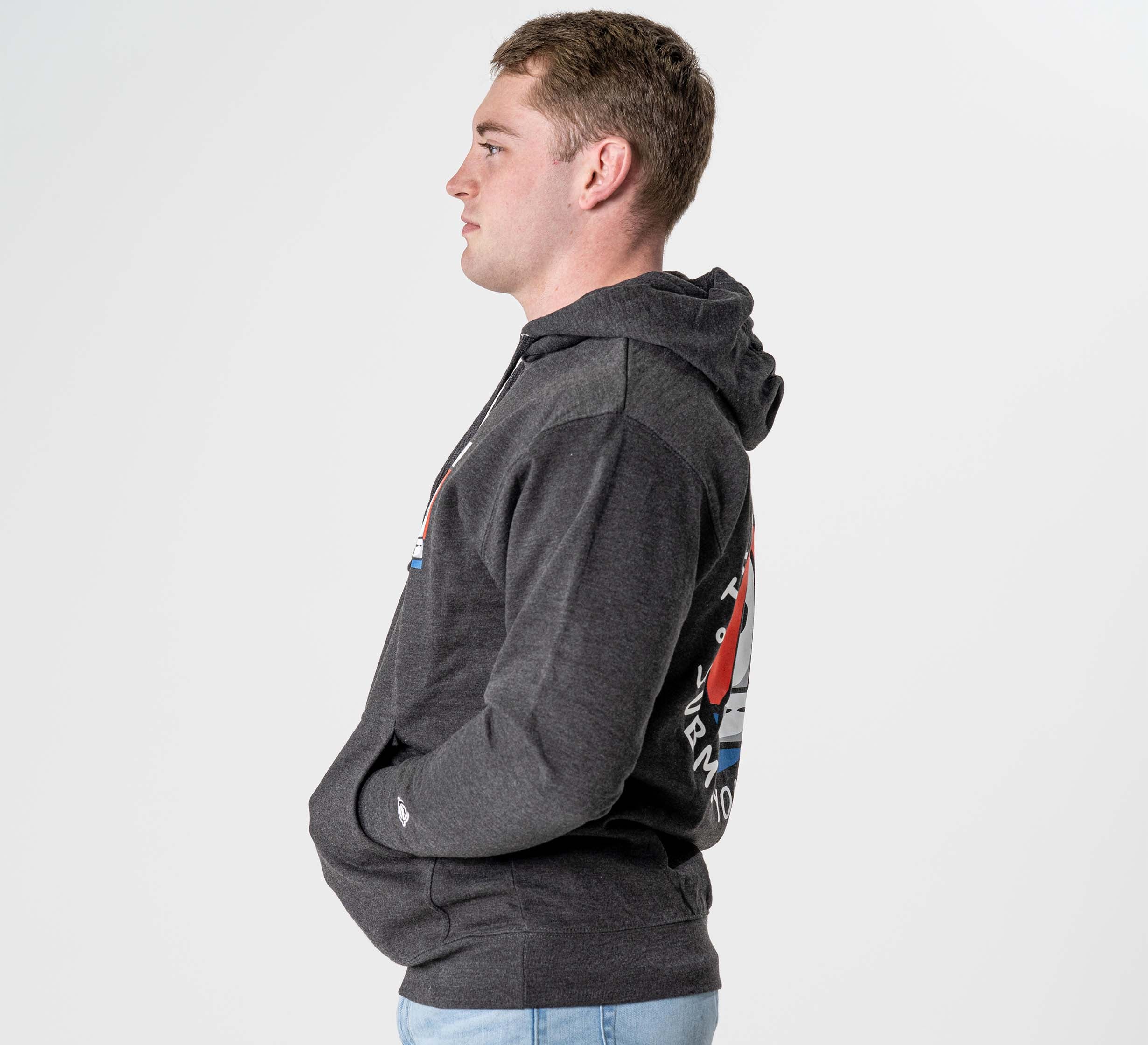 A young man stands in profile against a white background, wearing the Submit Your Ego Hoodie in heather black, featuring a colorful graphic on the back. He has his hands in the hoodie pockets and is dressed in light jeans.