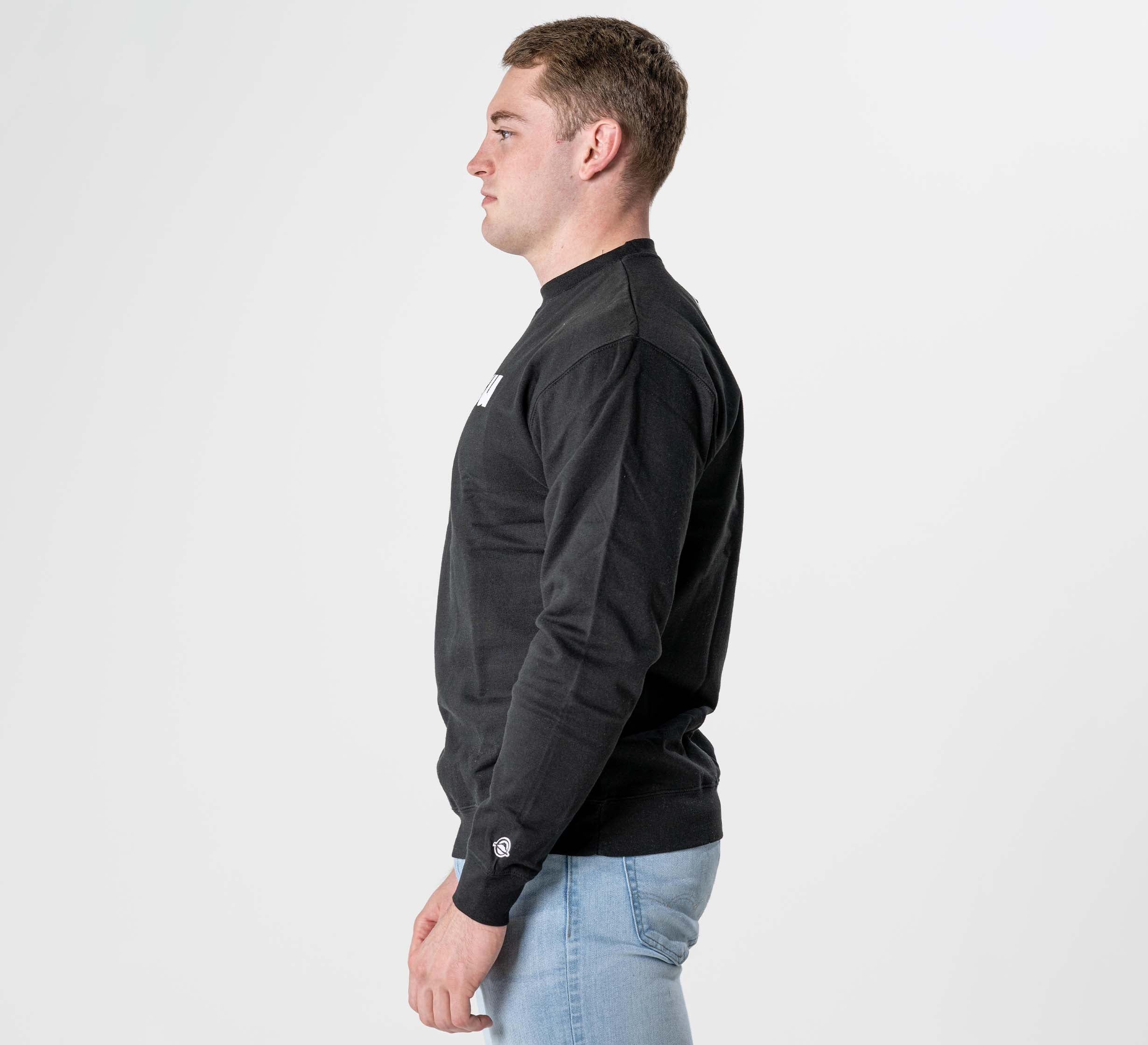 A man with short brown hair stands in profile, facing left, wearing the Jiu Jitsu Player Crewneck Black and light blue jeans against a plain white background.