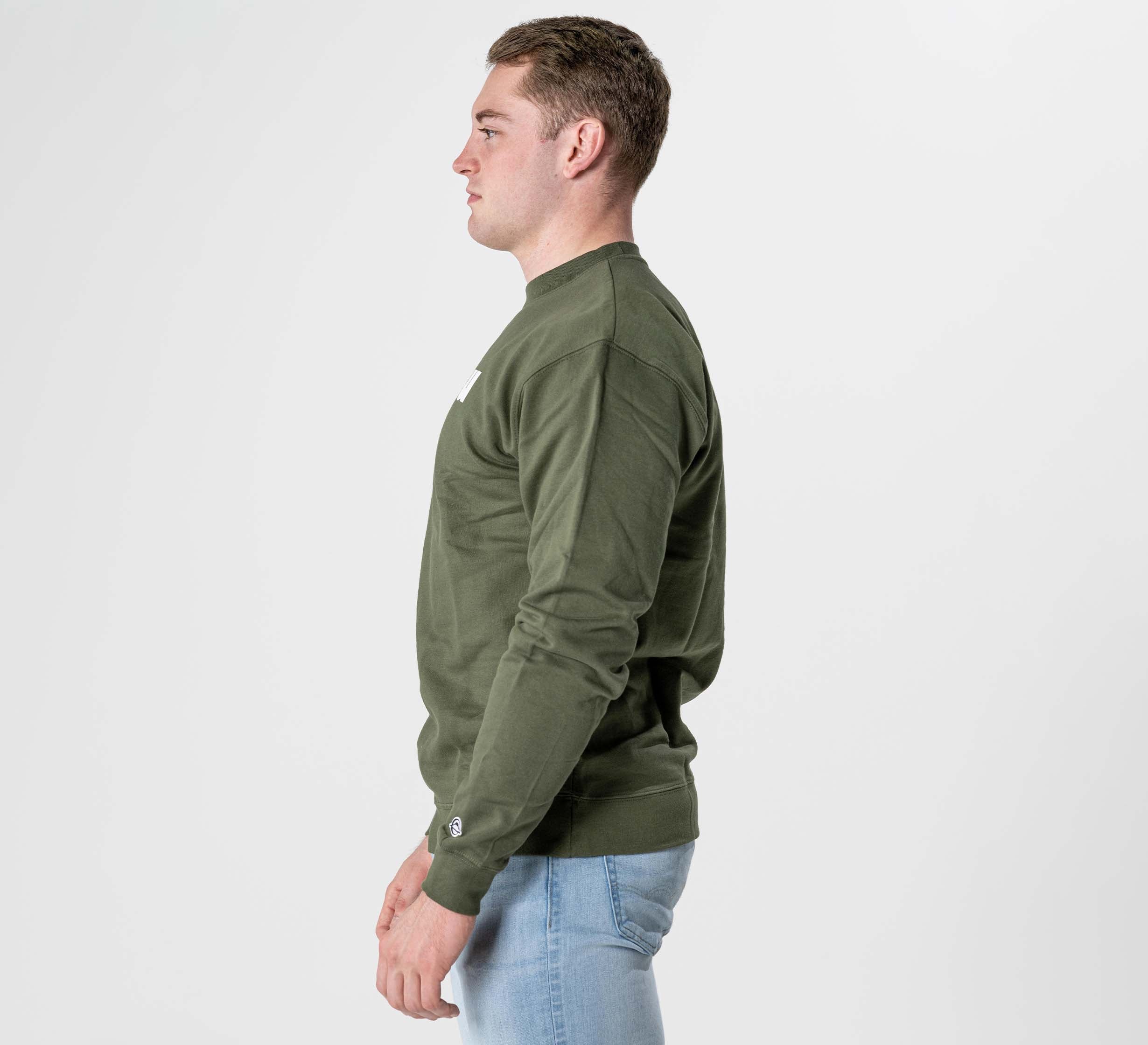 A man stands in profile facing left, wearing the Jiu Jitsu Player Crewneck Military Green and light blue jeans against a plain white background.
