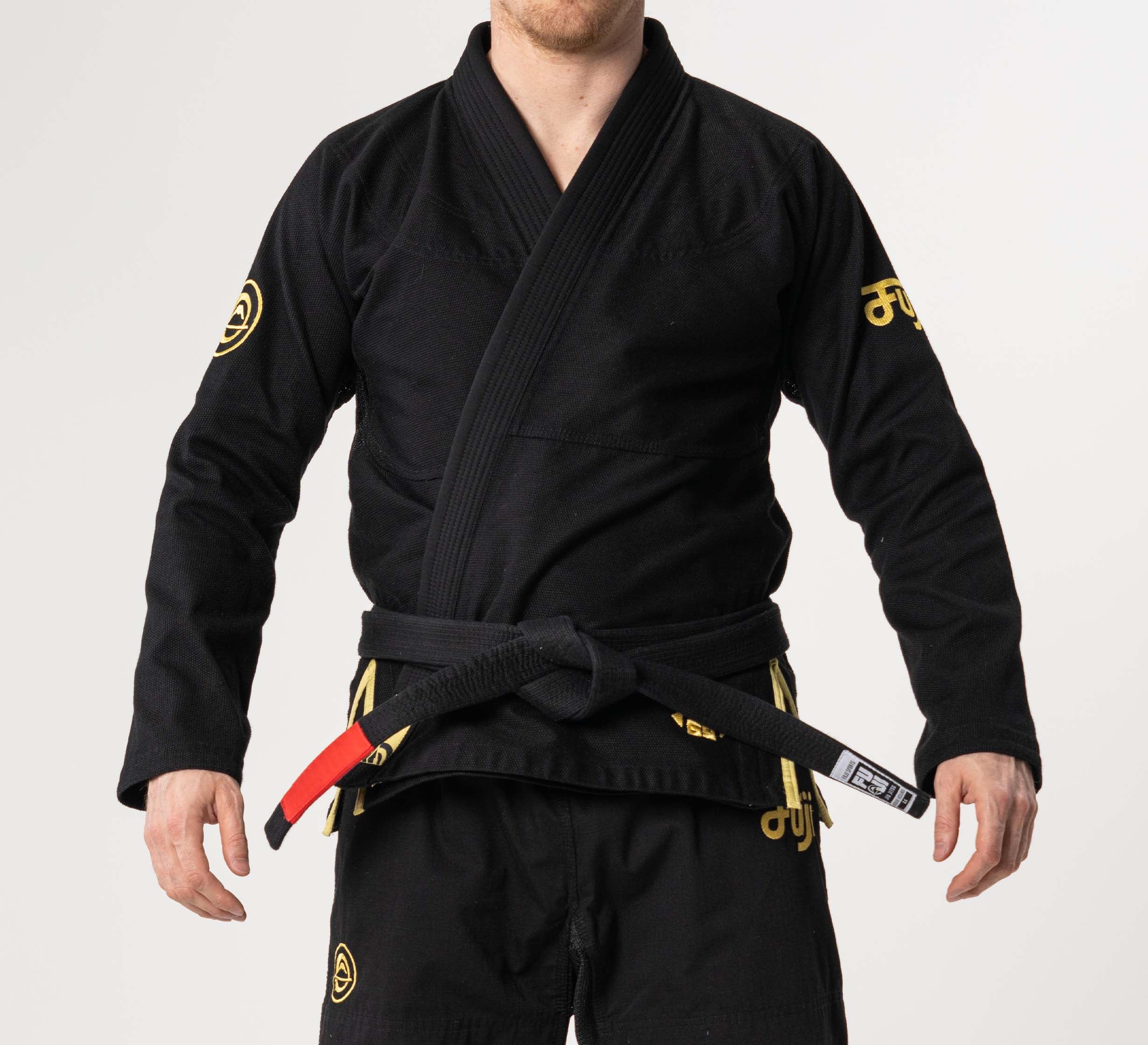 A person wearing the Flow-Tech BJJ Gi Black/Gold with yellow logos and a black belt with red bar, standing with arms slightly outstretched against a plain white background.