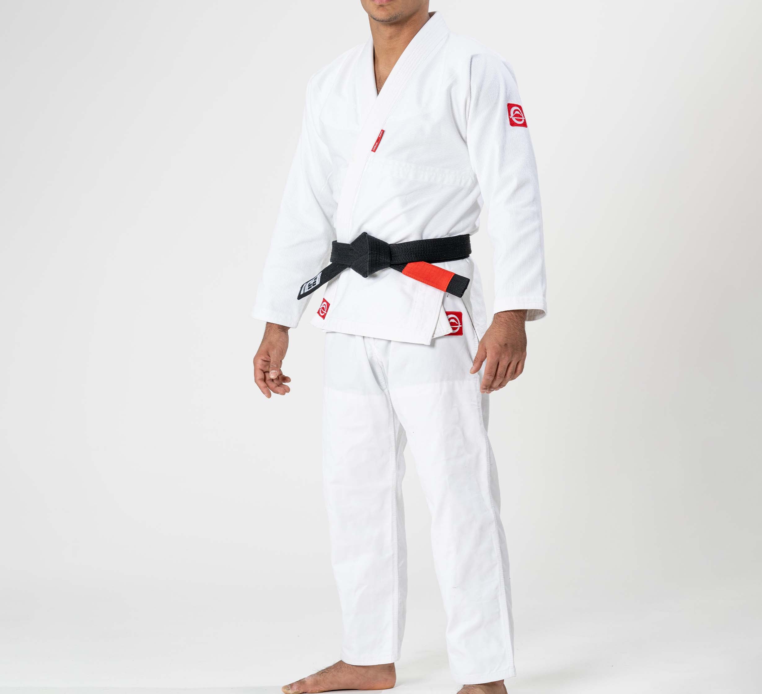 A person wearing the Ultra Lite 300 BJJ Gi White with a black belt, barefoot against a plain white background. The gi features red patches on the sleeves and waist.