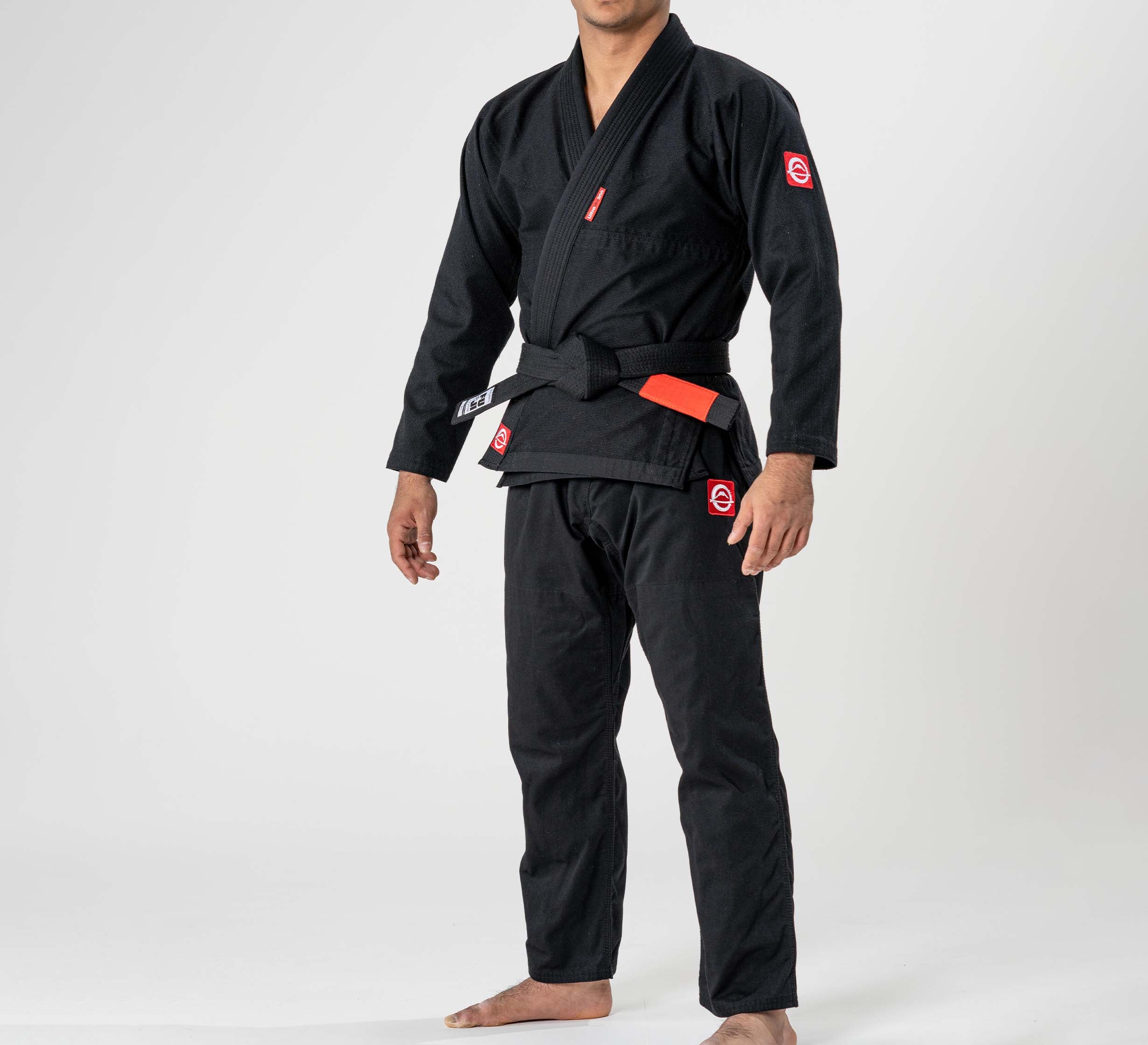 A child stands barefoot on a white background, wearing the Kids Ultra Lite 300 BJJ Gi Black with a black belt and red and white patches on the chest, sleeves, and pants. The child's face is not visible.
