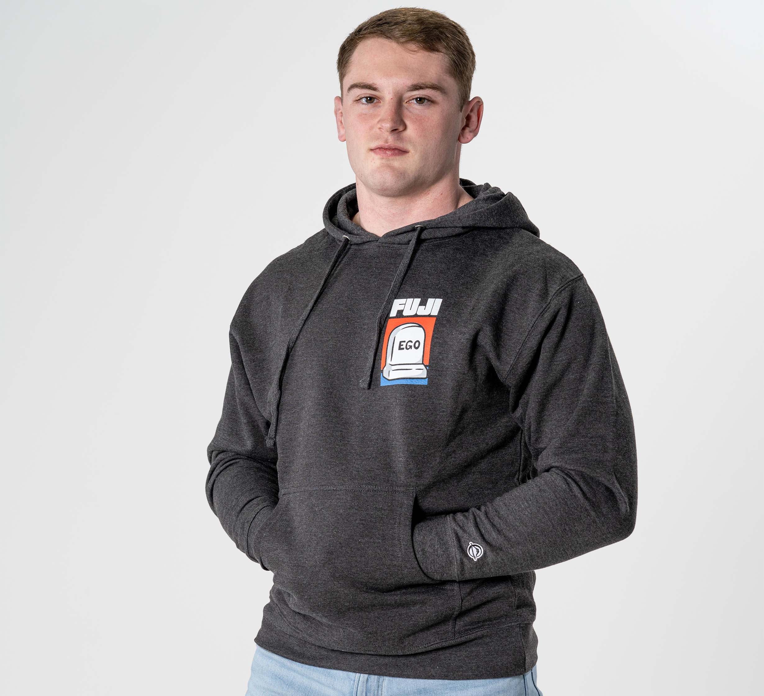 A young man stands against a plain white background, wearing the Submit Your Ego Hoodie in Heather Black with a vibrant FUJI EGO graphic on the chest, looking at the camera and keeping his hands in his pockets.