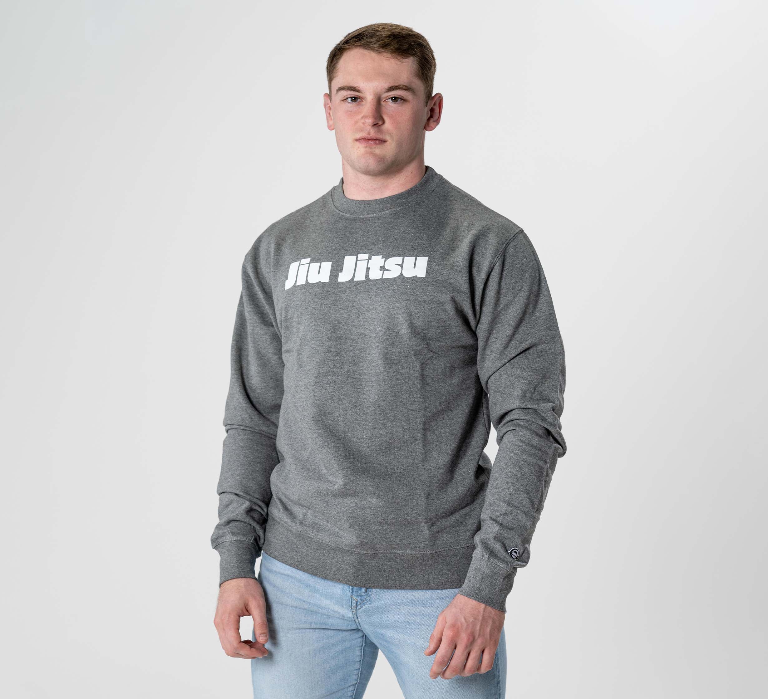 A young man faces the camera wearing the Jiu Jitsu Player Crewneck Heather Grey, featuring bold white "Jiu Jitsu" text on the chest, paired with light blue jeans against a plain light background.
