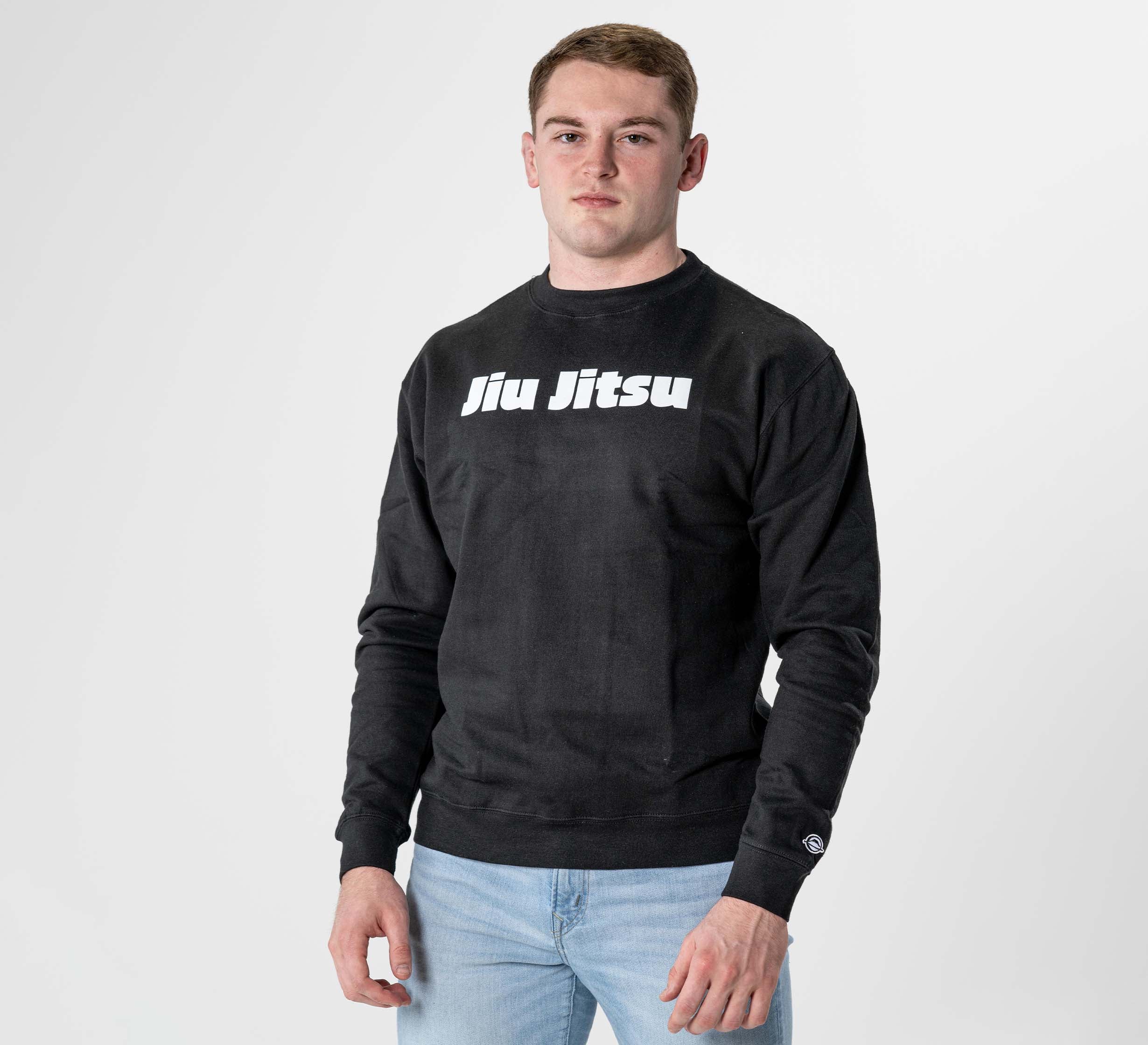 A man stands against a plain white background wearing the Jiu Jitsu Player Crewneck Black, featuring "Jiu Jitsu" in white letters across the chest, paired with light blue jeans.