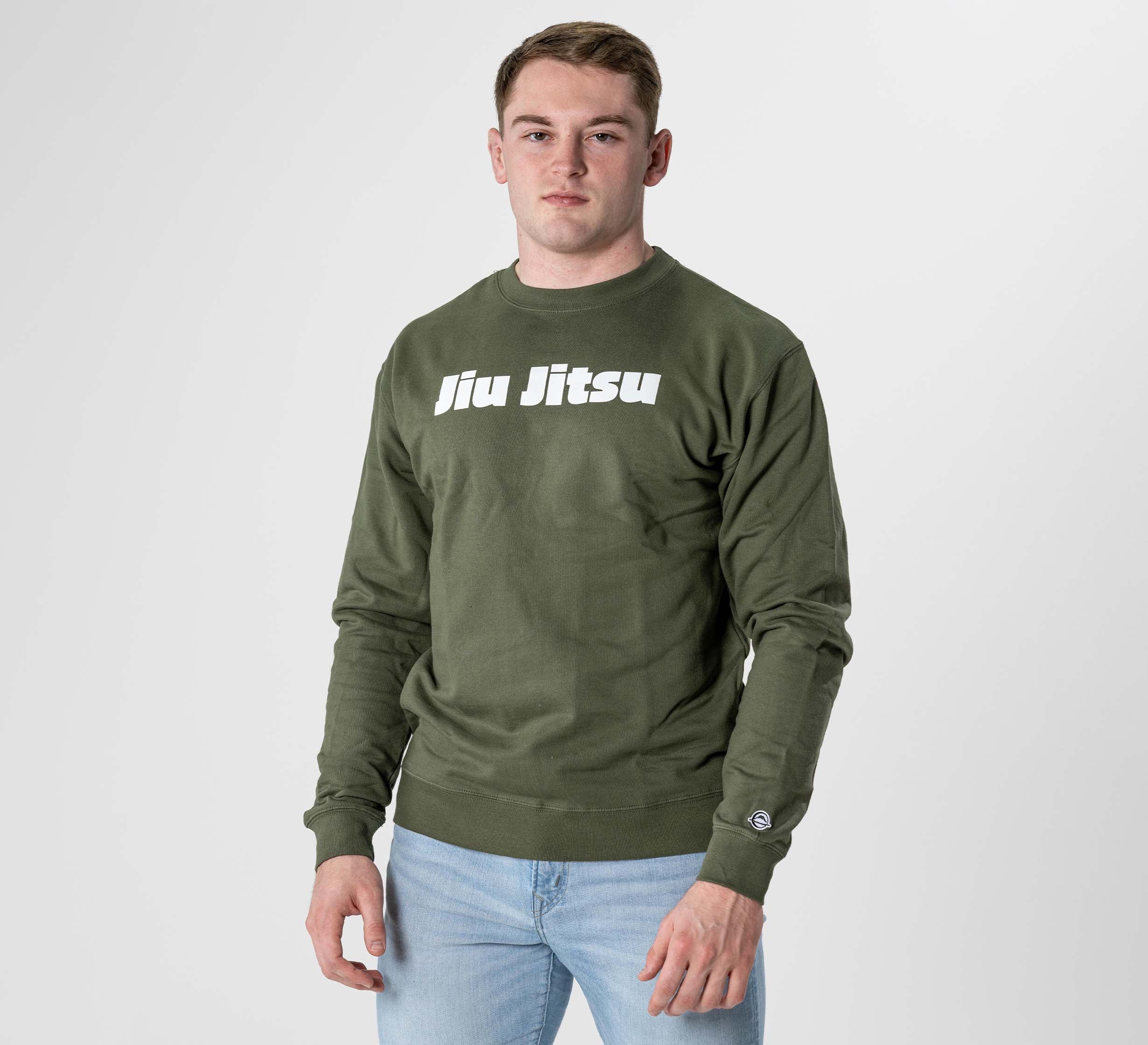 A young man stands against a plain white background, wearing the Jiu Jitsu Player Crewneck in military green with “Jiu Jitsu” printed in white on the front and light blue jeans.