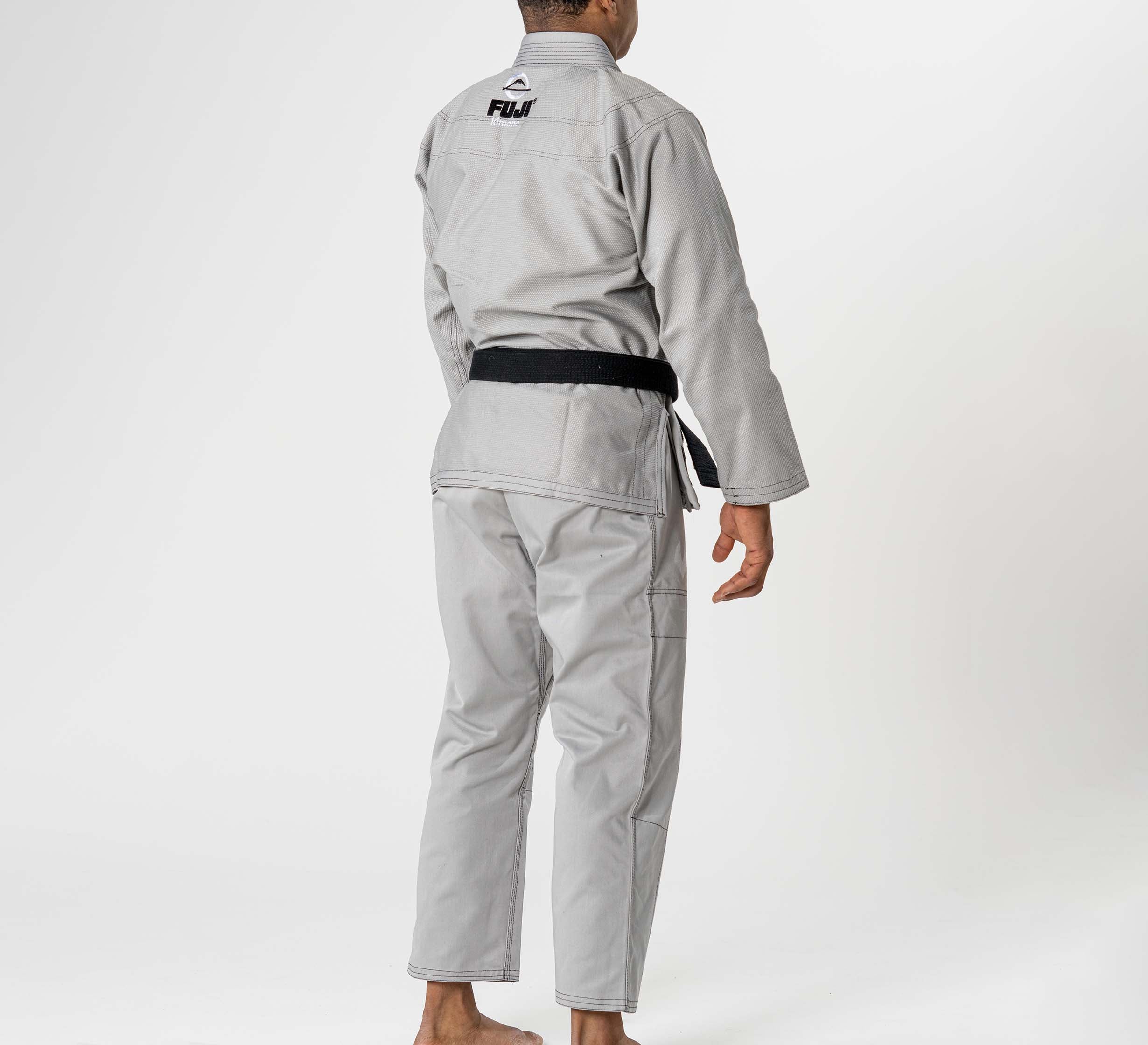 A person wearing the Lightweight BJJ Gi Grey with a black belt stands barefoot, facing away from the camera against a white background. The gi features "FUJI" and its logo on the upper back.