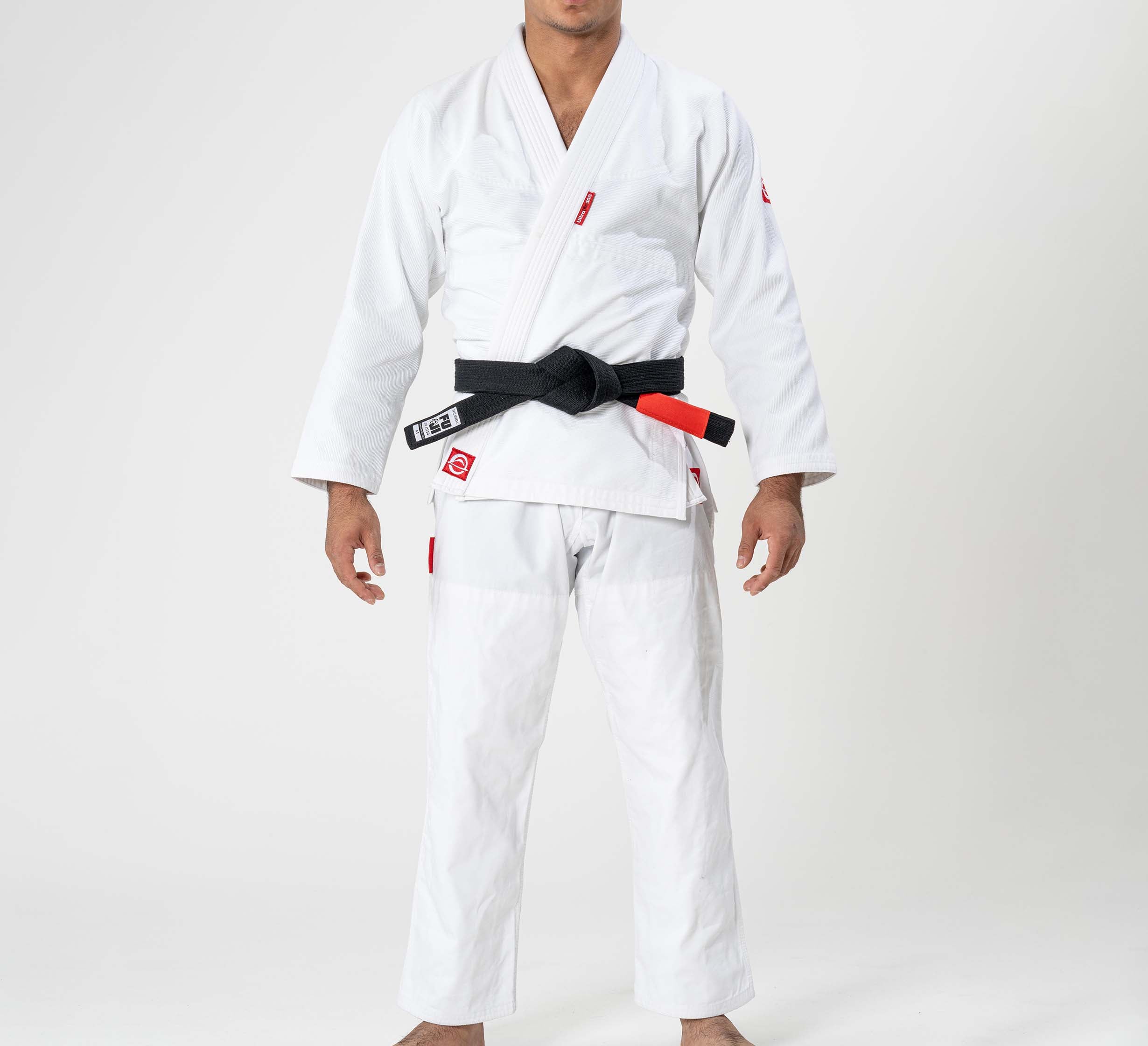 A child models the Kids Ultra Lite 300 BJJ Gi in white with a black belt, standing barefoot against a plain white background. Their face is not visible.