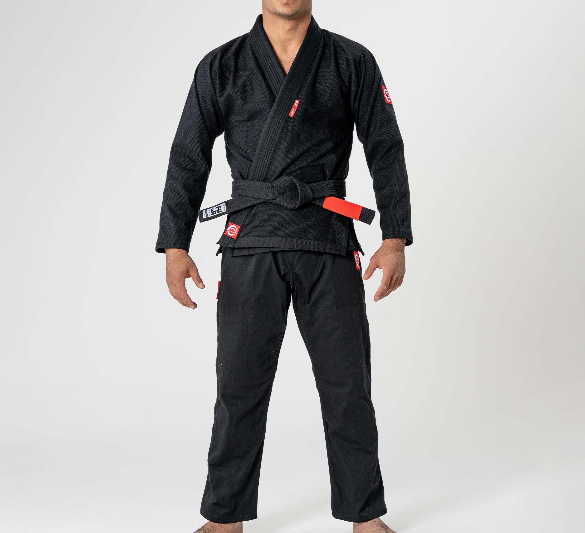 A child stands barefoot in a Kids Ultra Lite 300 BJJ Gi Black with a black belt, facing forward against a plain white background. Their head is partially out of frame.
