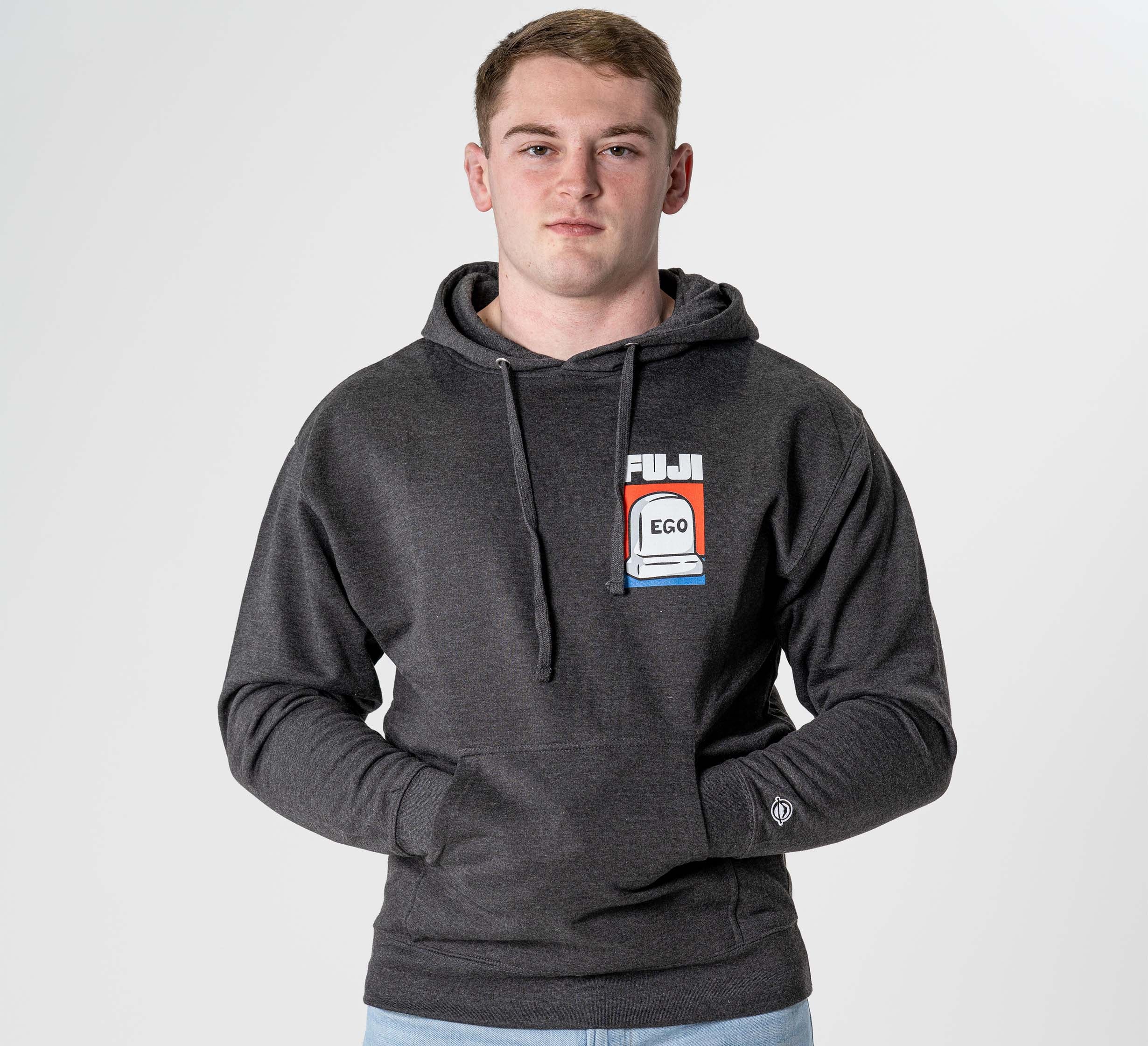 A young man wears the Submit Your Ego Hoodie in Heather Black, standing against a plain white background, looking directly at the camera with a neutral expression and his hands together.