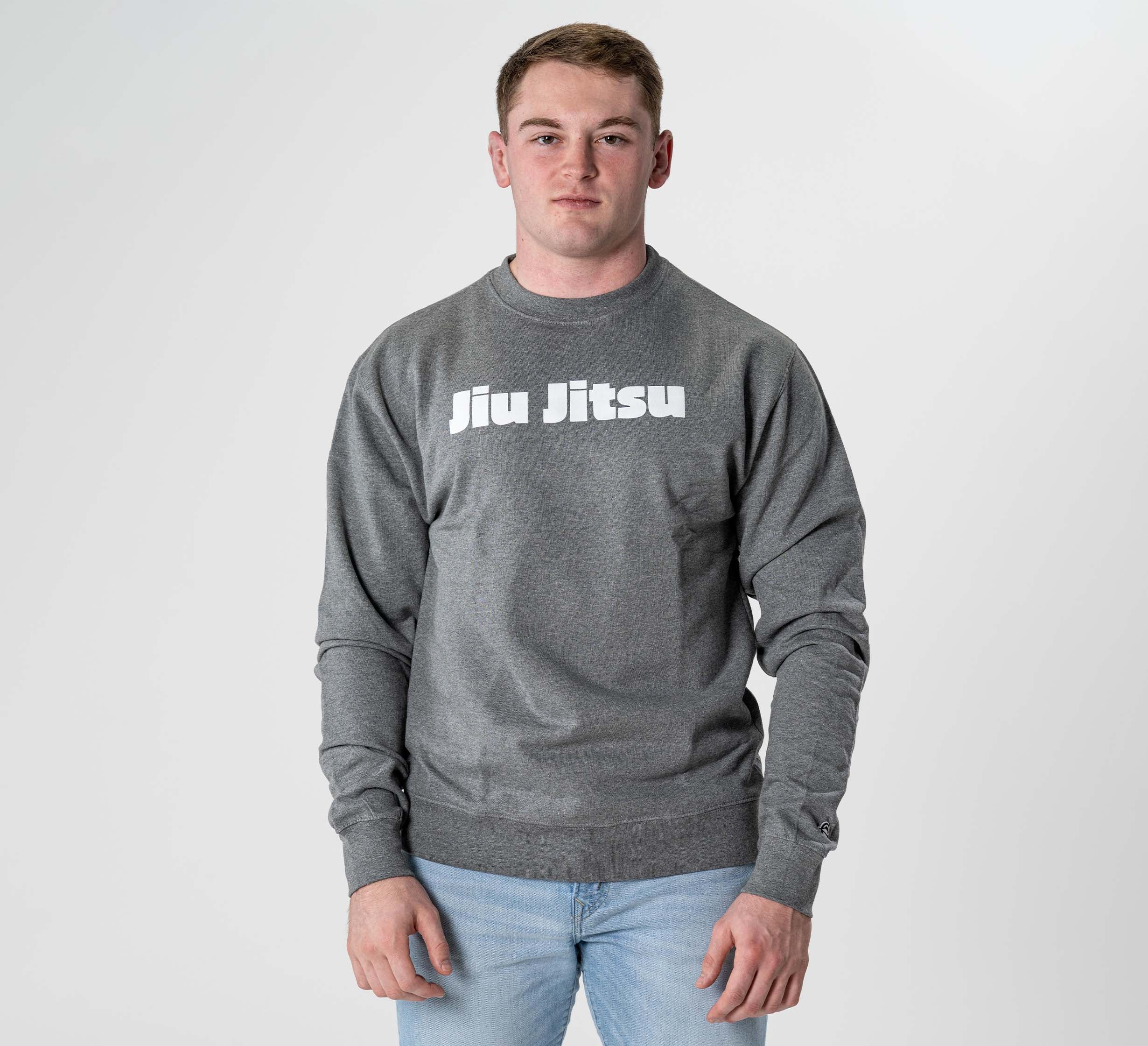 A young man with short hair stands against a light background wearing the Jiu Jitsu Player Crewneck Heather Grey, which features "Jiu Jitsu" in white print. He looks directly at the camera with a neutral expression.