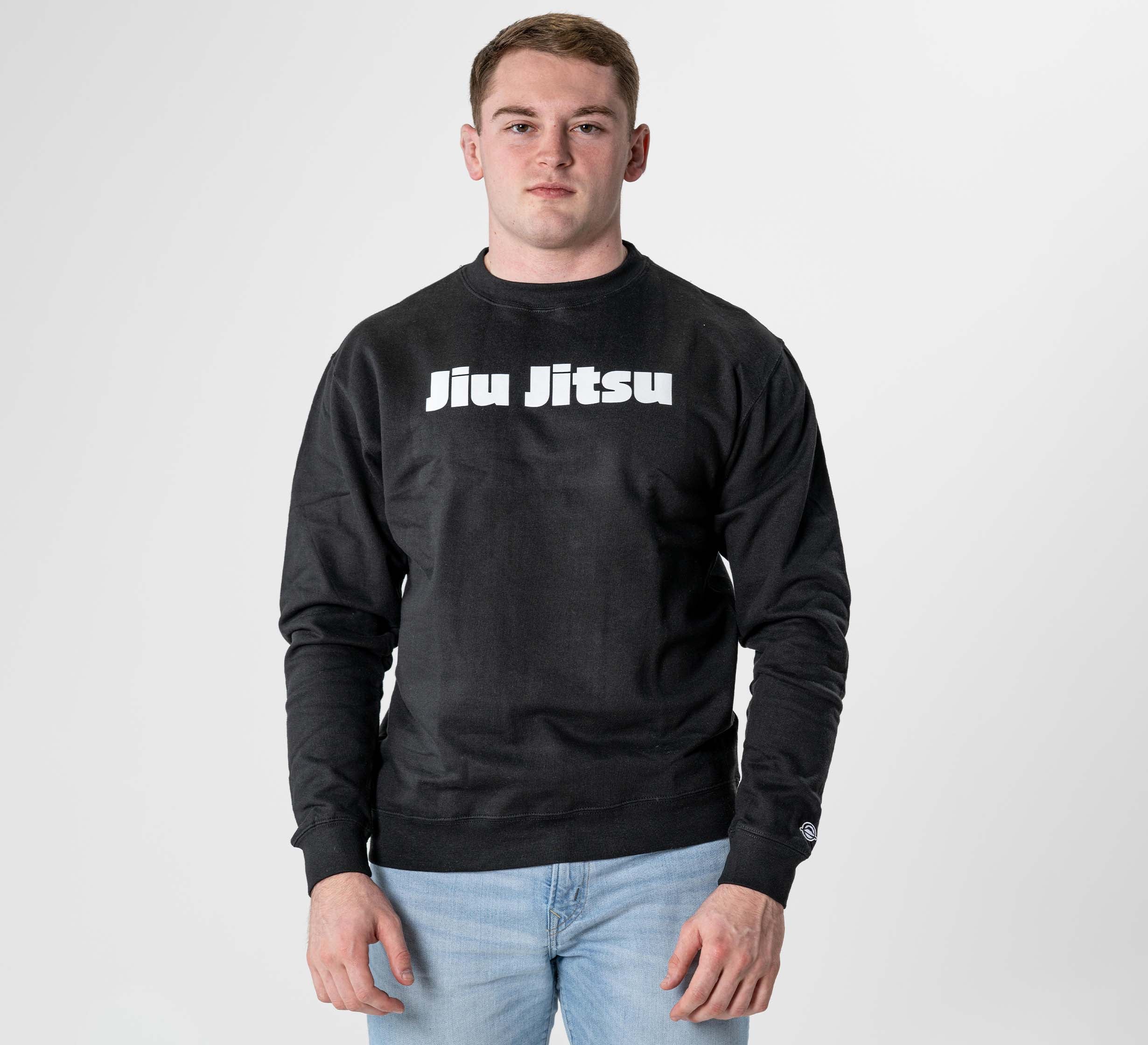 A man faces forward in the Jiu Jitsu Player Crewneck Black, featuring bold white "Jiu Jitsu" lettering on the front, paired with light blue jeans against a plain white background.