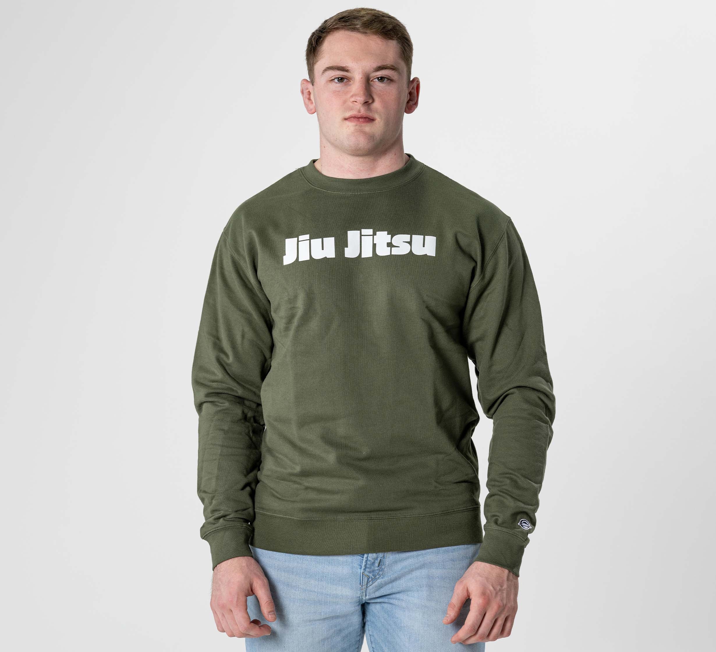 A young man stands against a plain light background, wearing the Jiu Jitsu Player Crewneck in military green with bold white "Jiu Jitsu" lettering on the chest, paired with light blue jeans.
