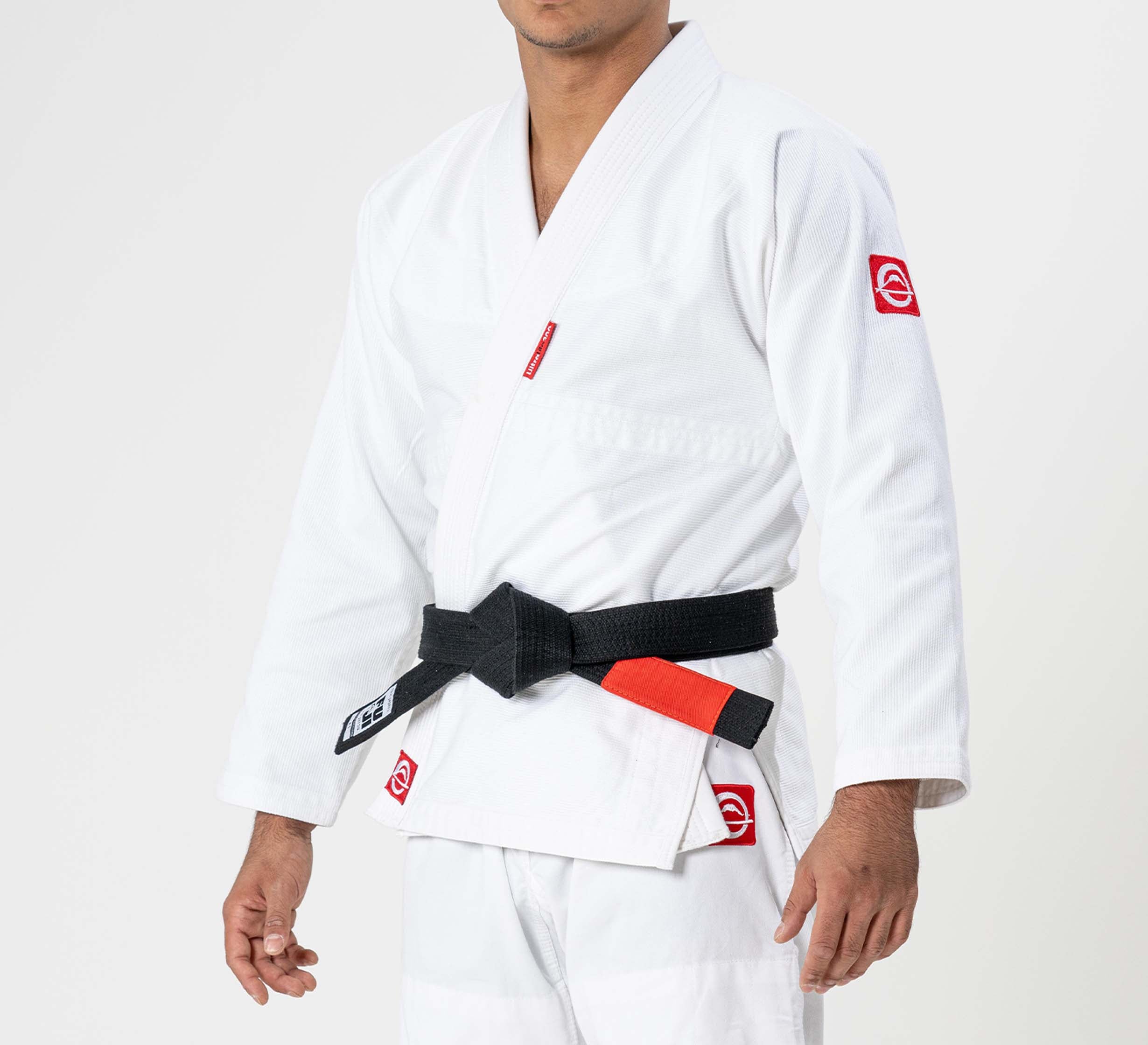 A child wears the Kids Ultra Lite 300 BJJ Gi in white, featuring red sleeve patches and a black belt with red bar, standing against a plain light background with their face not visible.