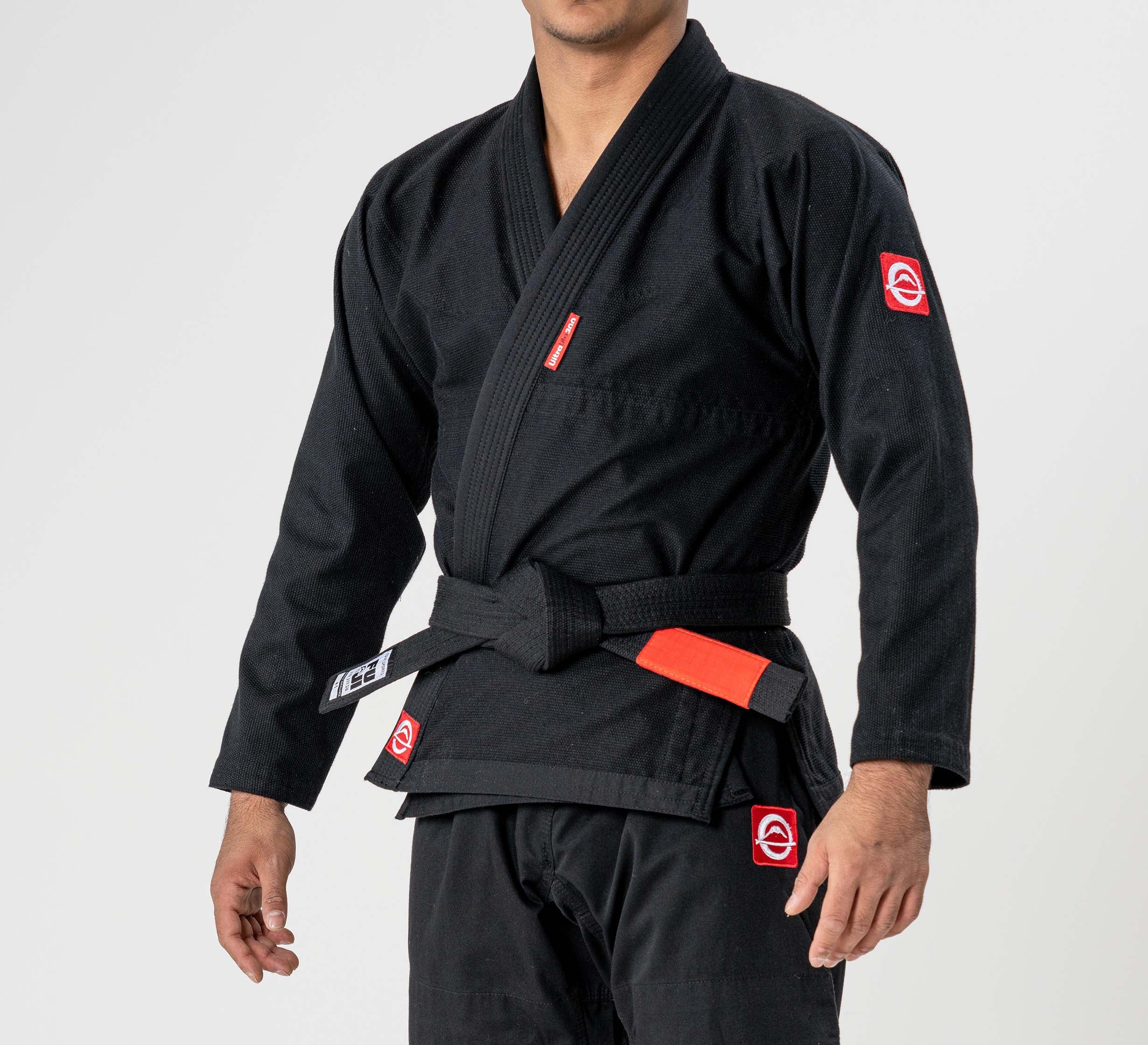 A person wearing an Ultra Lite 300 BJJ Gi Black with red and white patches, paired with a black belt featuring a red bar, stands against a plain light background.