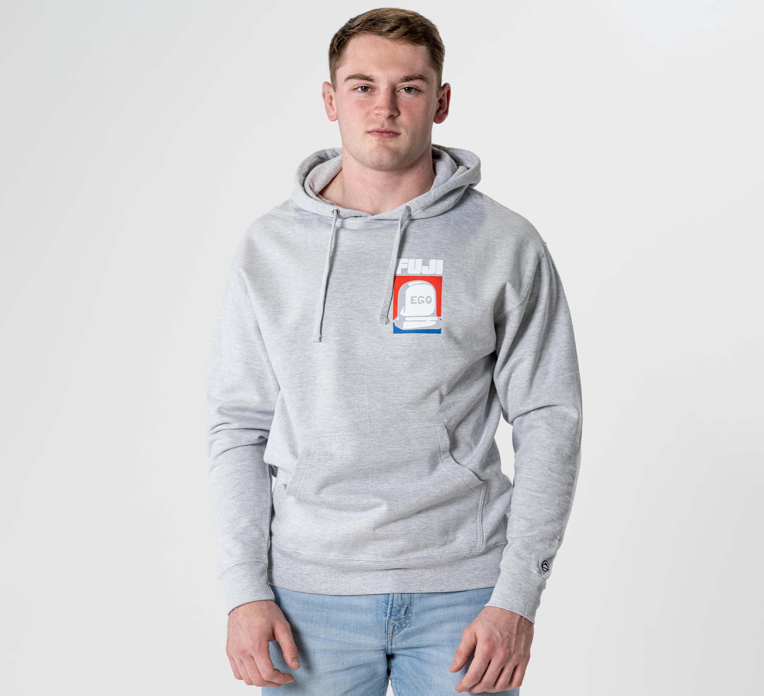 A young man with short brown hair wears the Submit Your Ego Hoodie in heather grey, featuring a colorful chest graphic, paired with light blue jeans, standing against a plain white background.