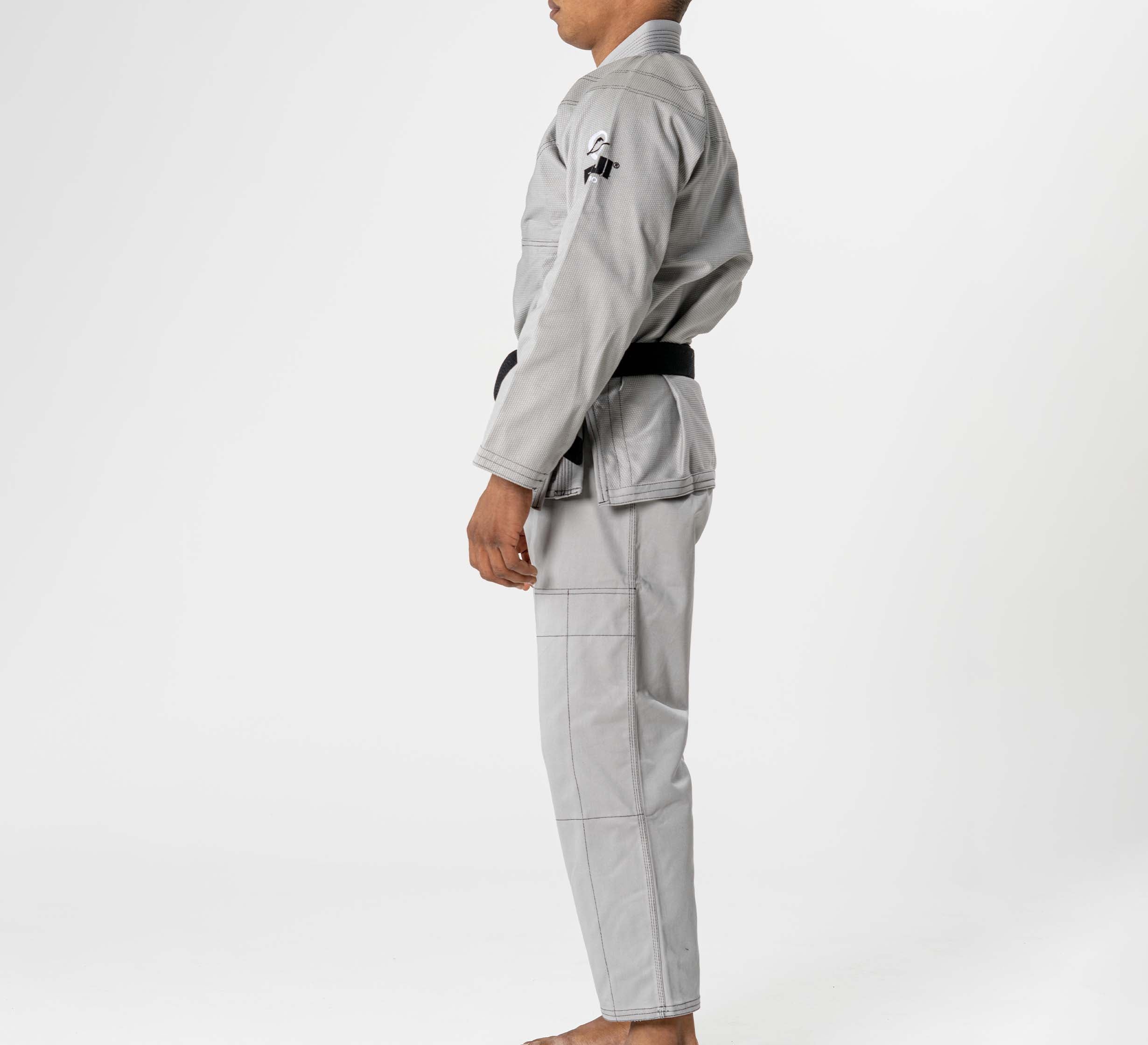 A person stands in profile, barefoot, against a plain white background, wearing the Lightweight BJJ Gi Grey and a black belt.