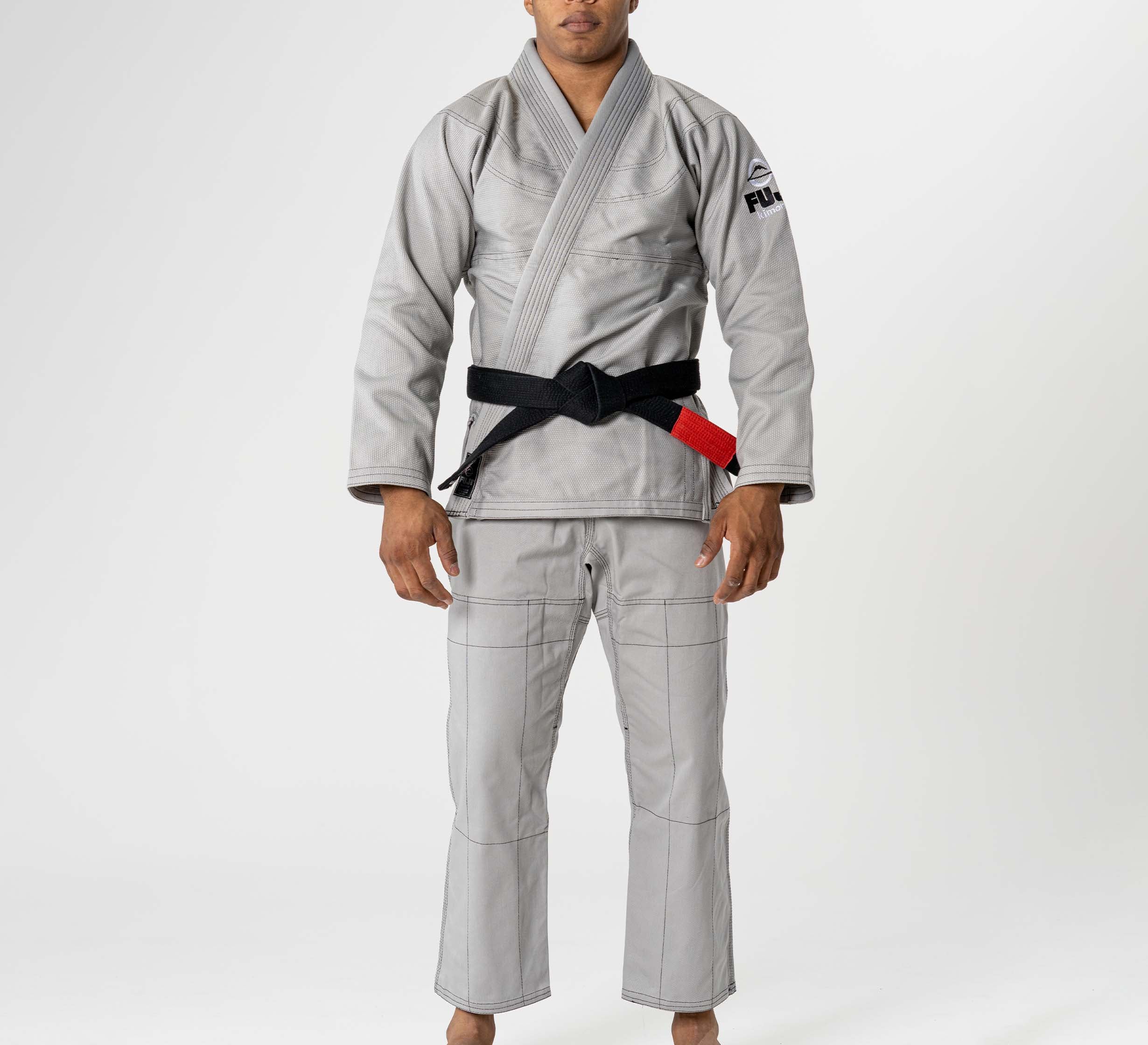 A person wearing the Lightweight BJJ Gi Grey with visible stitching and a patch on the upper sleeve, along with a black belt, stands barefoot against a plain white background.