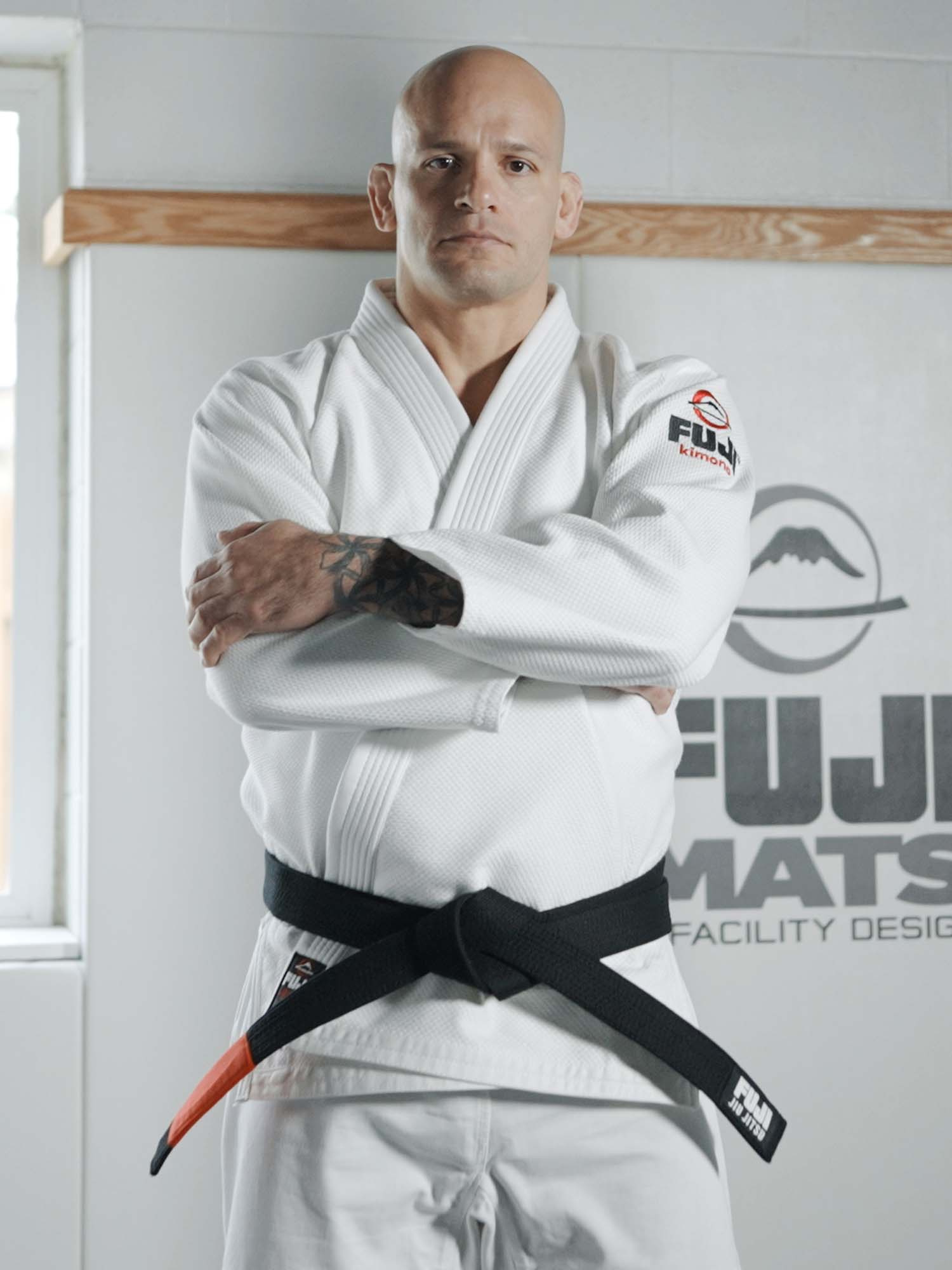 A man in the All Around BJJ Gi White with a black belt stands with arms crossed before a wall displaying the Fuji Mats logo.