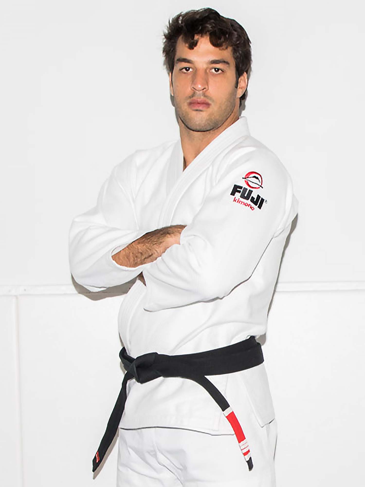 A man stands confidently with arms crossed, wearing the All Around BJJ Gi White with a black belt and FUJI logo, against a plain white background.