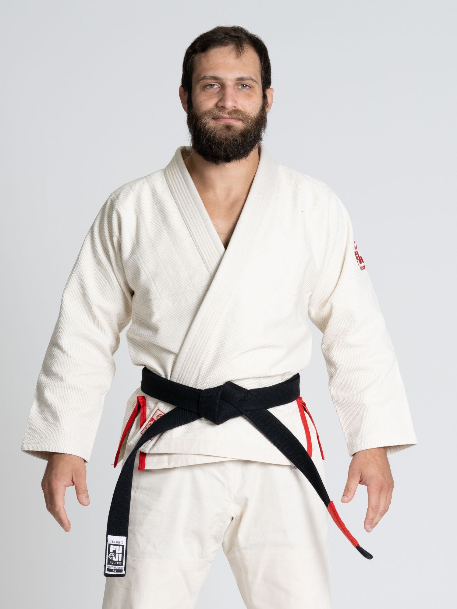A bearded man stands facing forward against a plain light background, smiling slightly with his hands relaxed at his sides, wearing the All Around BJJ Gi Natural and a black belt.