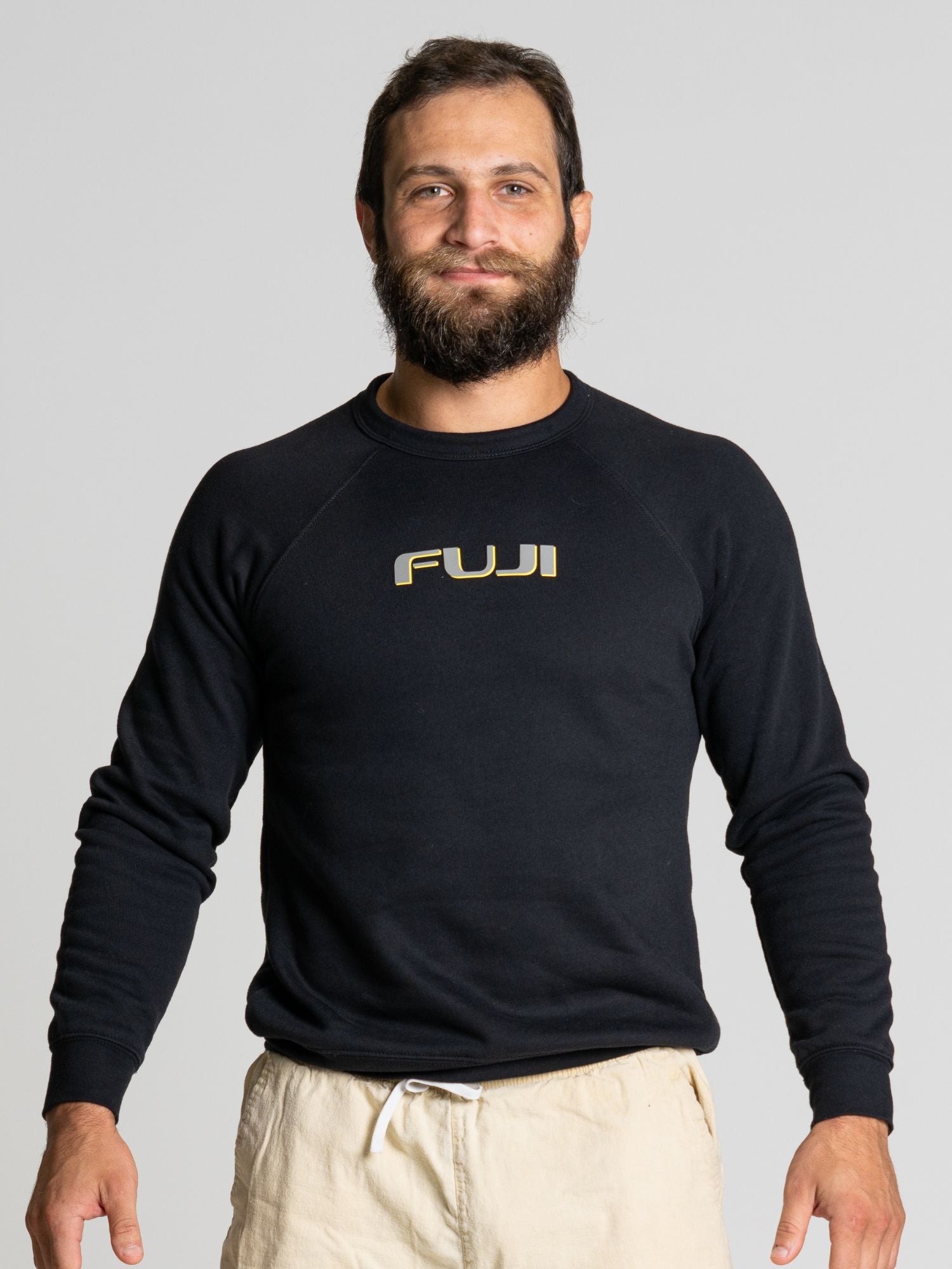 A man with a beard and mustache is smiling, wearing the Oni Legend Crewneck Black sweatshirt and light-colored pants, standing against a plain light gray background.