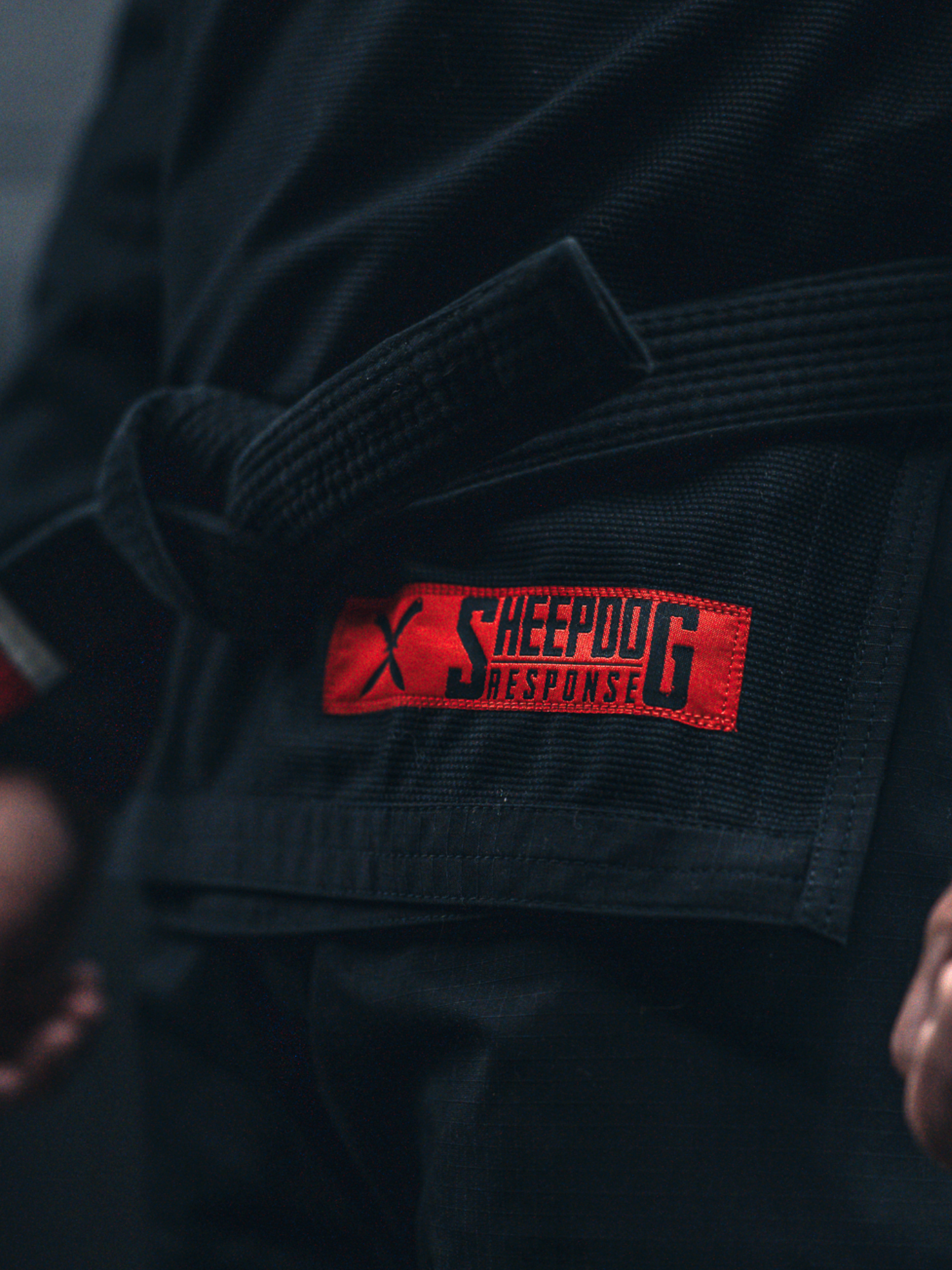 Close-up of a person wearing the Sheepdog Response Standard Suparaito BJJ Gi Black, featuring a black belt and a red “Sheepdog Response” patch on the gi. The person’s hands are visible at their sides.