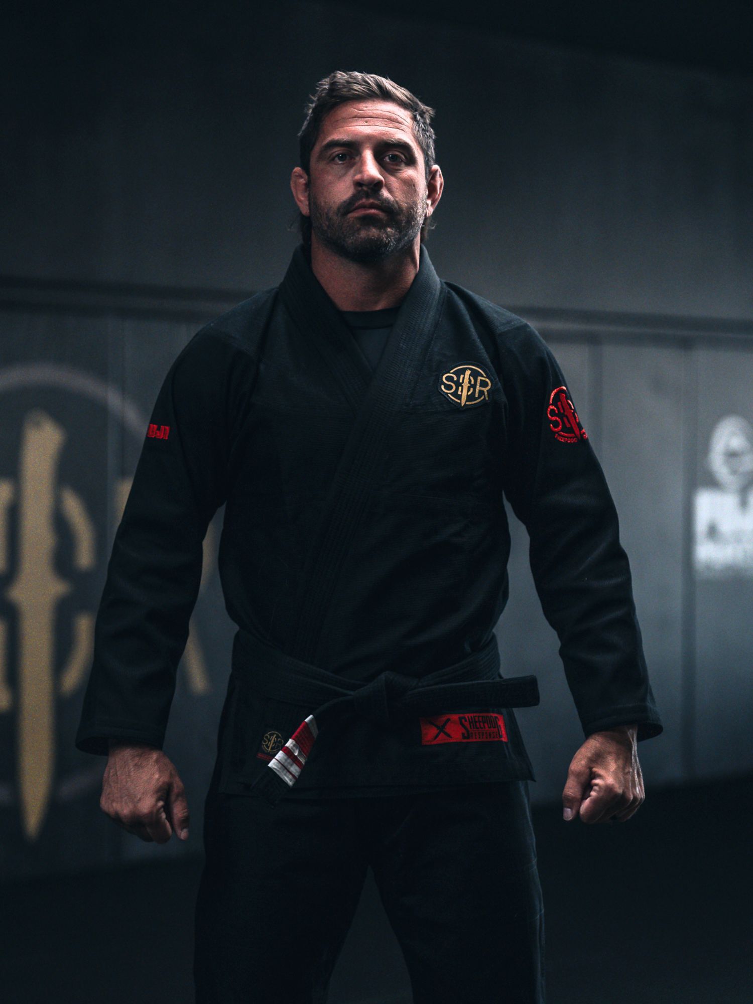 A man stands confidently in a dimly-lit gym, wearing the Sheepdog Response Standard Suparaito BJJ Gi Black adorned with red and yellow patches, his expression serious.
