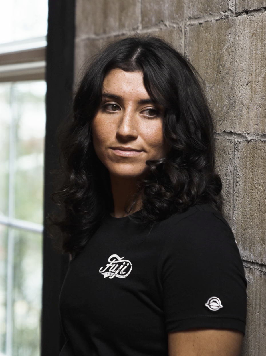 A woman with long, wavy dark hair and medium skin tone stands against a textured brick wall, wearing a black t-shirt with white logos. She gazes slightly to the side with a neutral expression. A window is visible in the background.