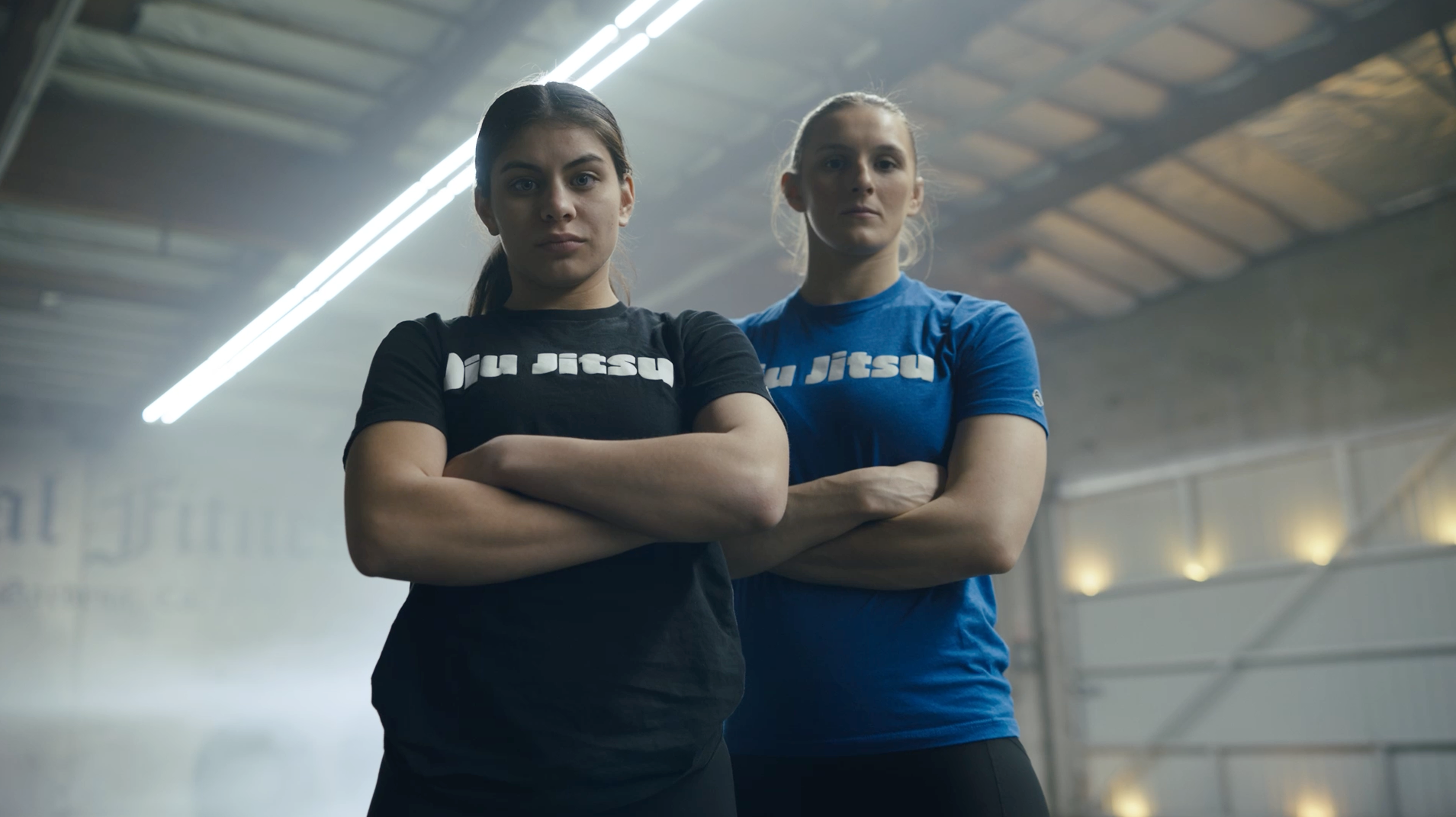 Two women stand confidently with arms crossed in a gym, wearing Jiu Jitsu t-shirts—one in black and the other in blue. Industrial lights and a high ceiling are visible in the background.