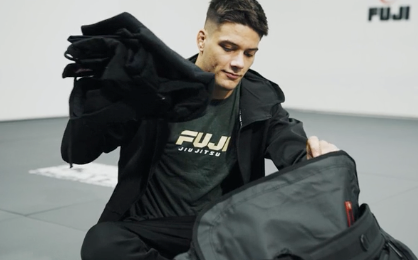 A person in a FUJI Jiu Jitsu shirt sits on a mat, holding a black garment in one hand and packing or unpacking a gym bag with the other. The FUJI logo is visible on the wall behind them.