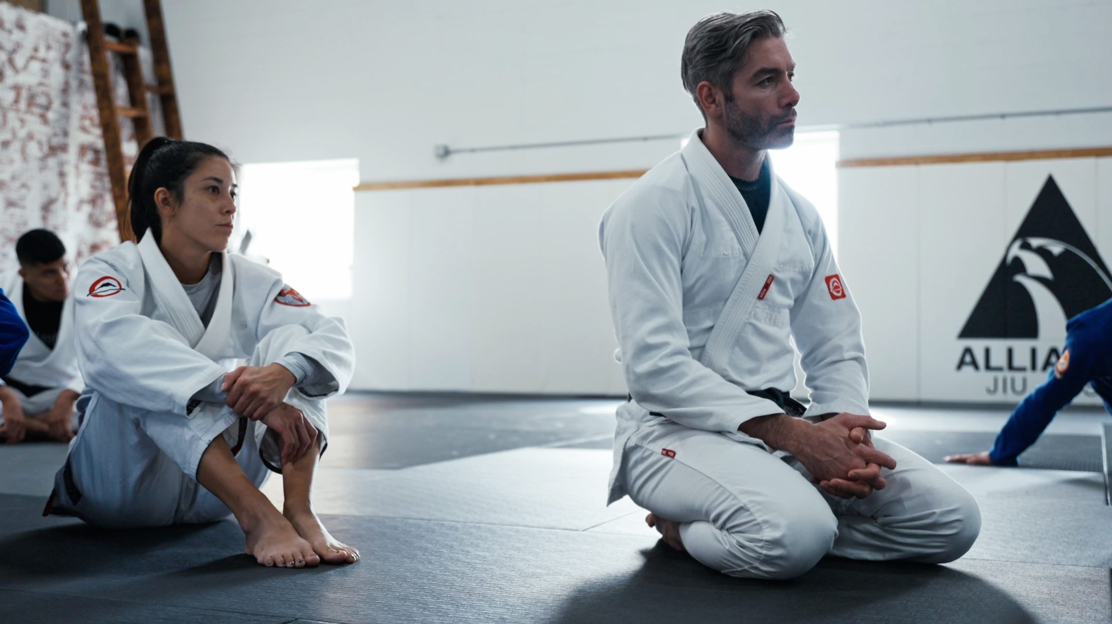Two people in white martial arts uniforms sit on the mat in a brightly lit dojo, listening attentively. One sits cross-legged while the other kneels. A wall with the word ALLIANCE is visible in the background.