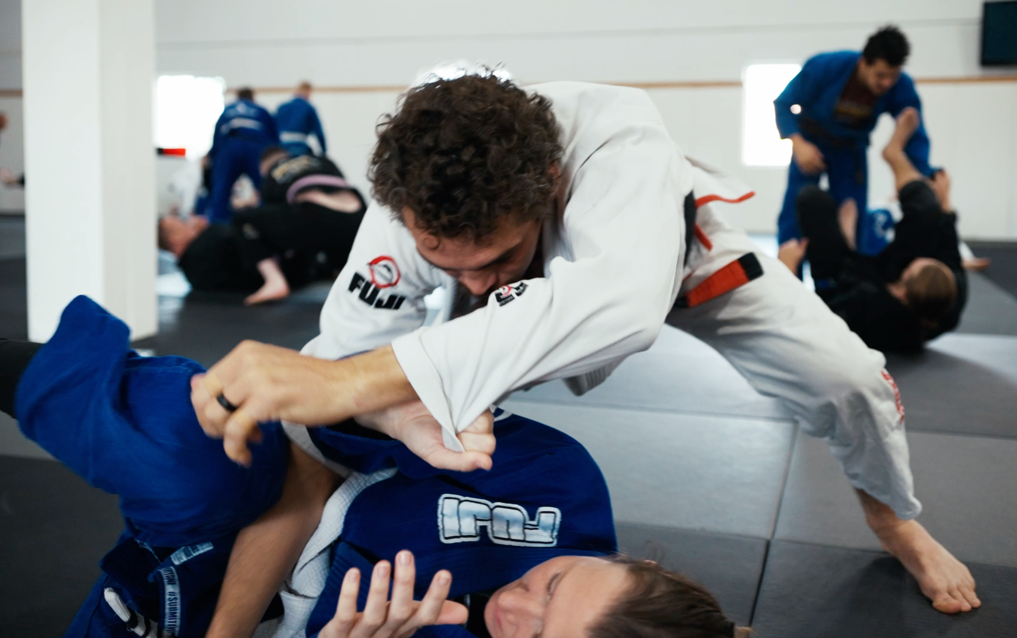 Two people wearing martial arts uniforms practice Brazilian Jiu-Jitsu on a mat. One person in a white gi is grappling and reaching over a person in a blue gi lying on their back. Others train in the background.
